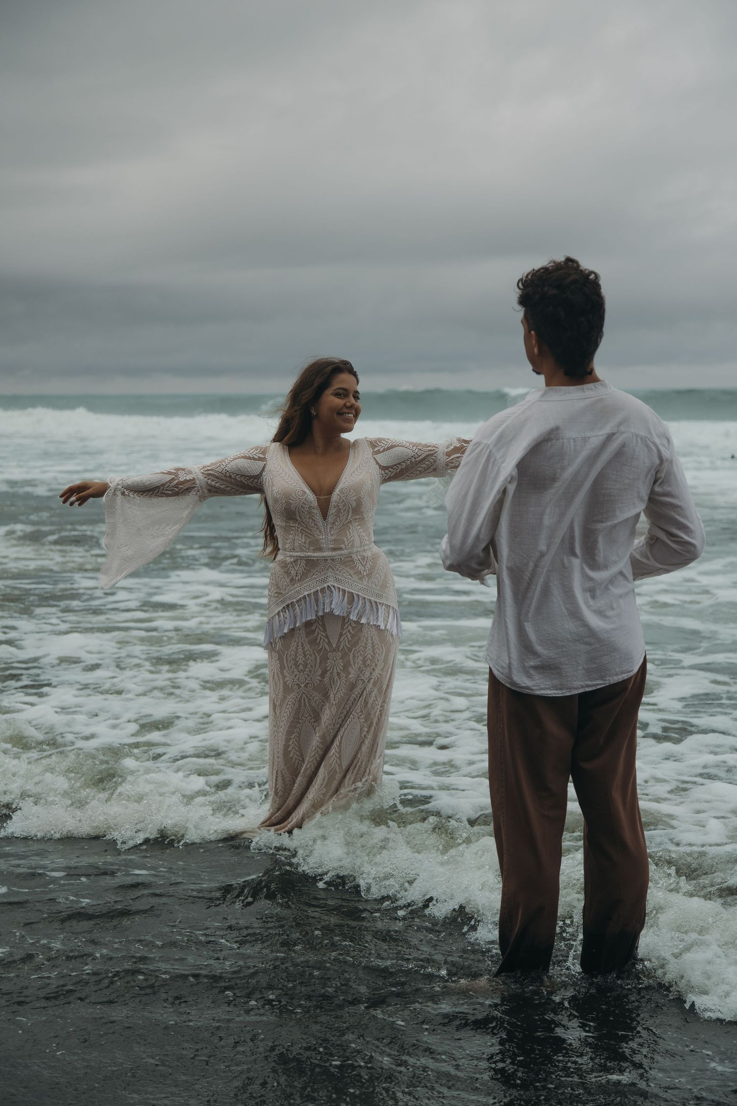 Joyful bride in wet lace dress spreads arms in ocean waves as groom watches on cloudy beach.