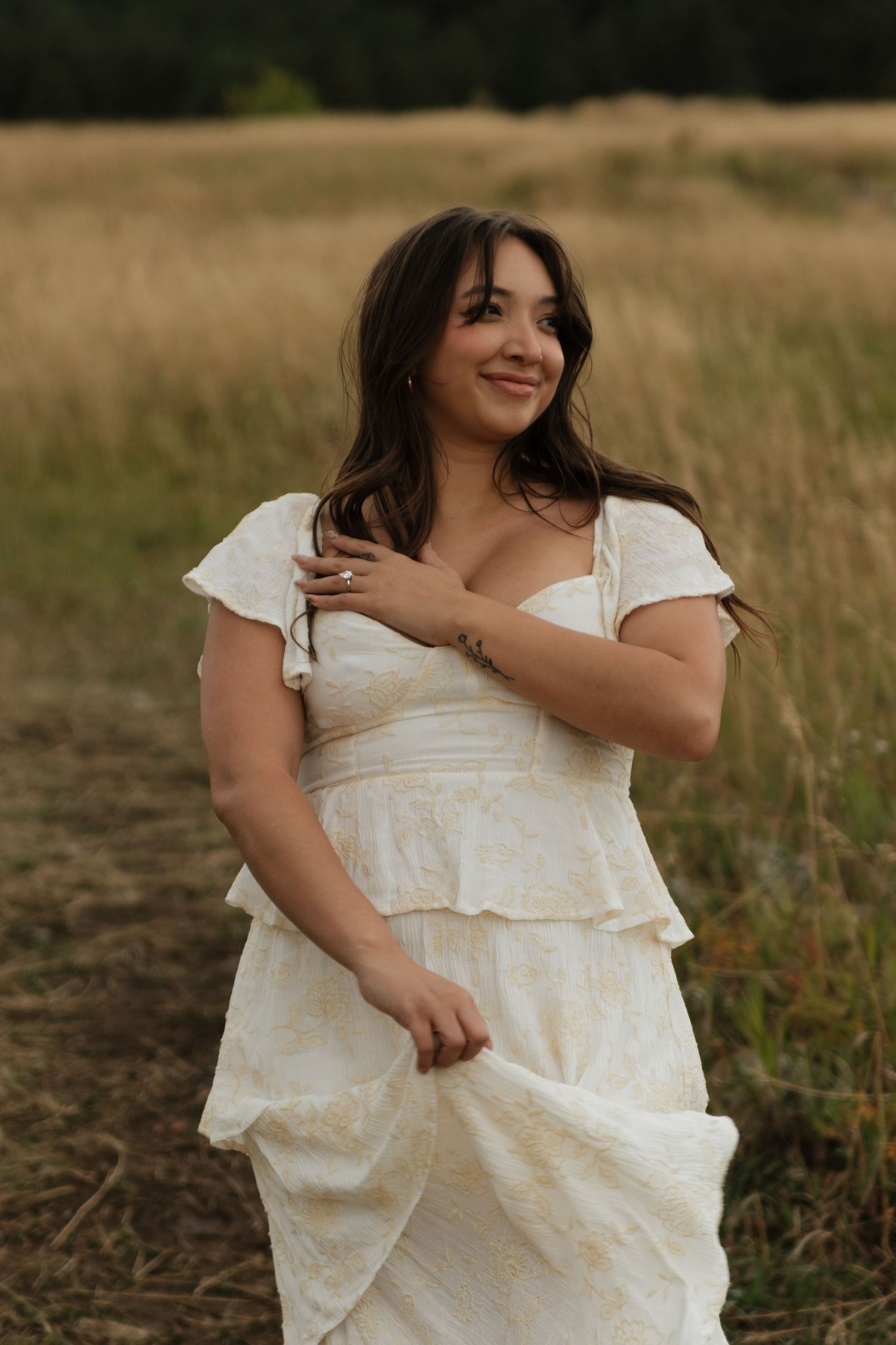 Woman in white tiered dress smiling in golden grass field, hand resting on chest.