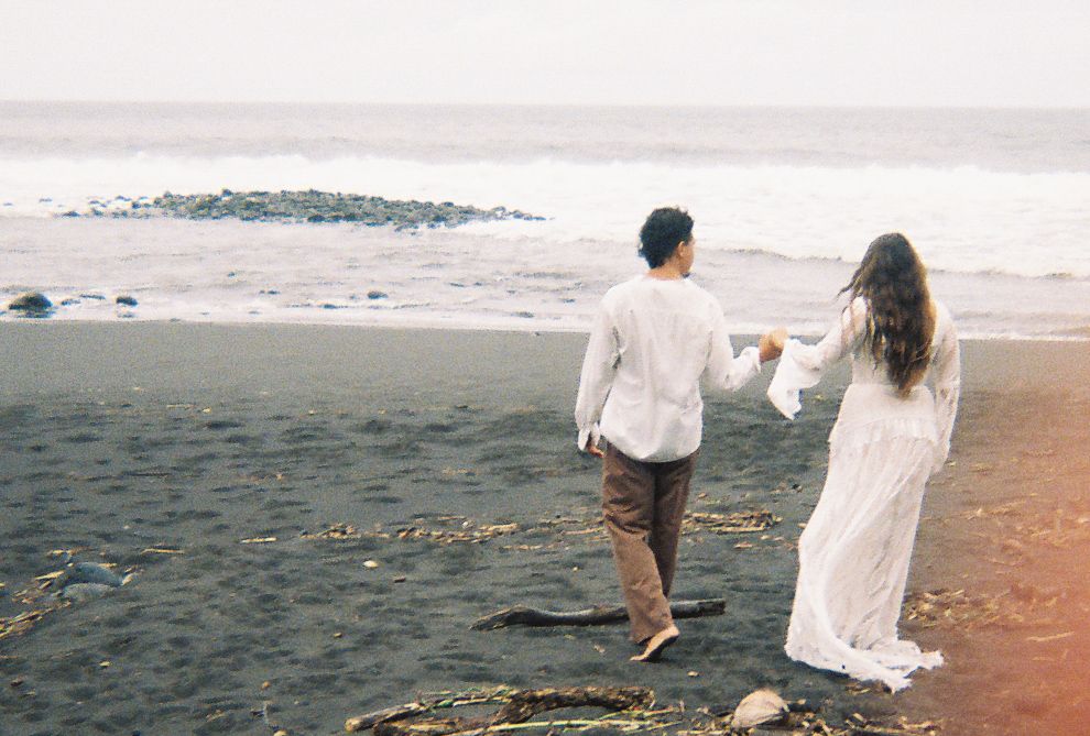 Couple walking hand in hand on a black sand beach toward the ocean, dressed in white and tan romantic attire.