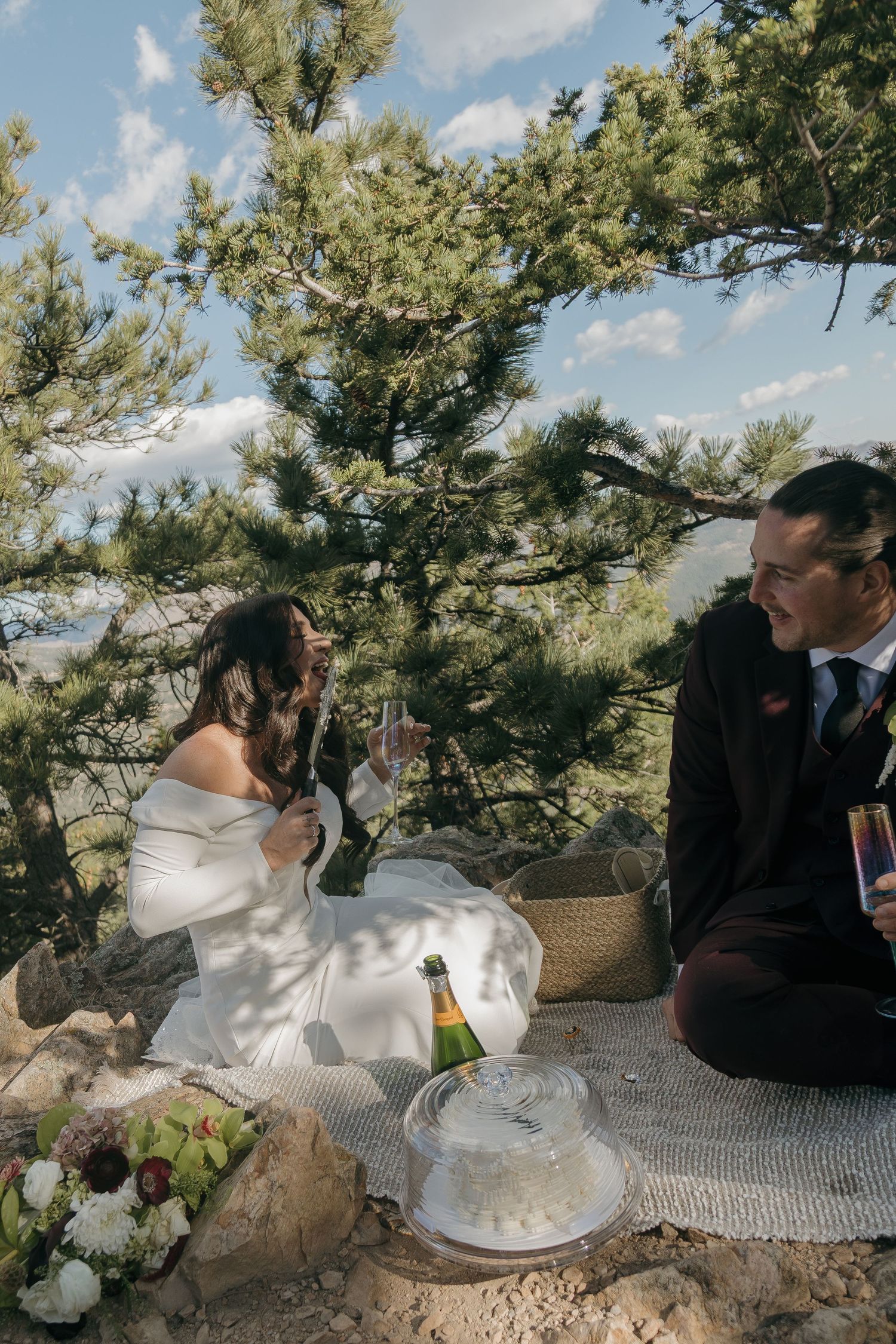 Bride and groom enjoying a champagne picnic under pine trees on a mountain rocky ledge after elopement.