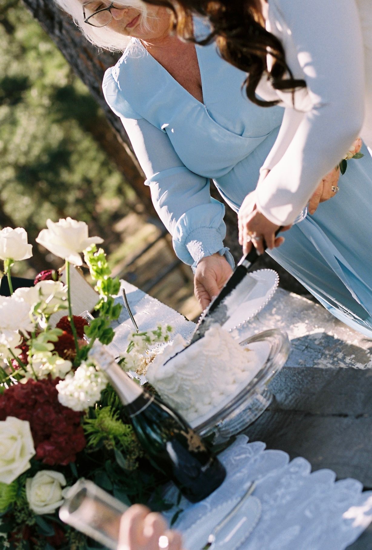 Bride in white gown cuts wedding cake beside lush floral arrangement of white and red flowers.