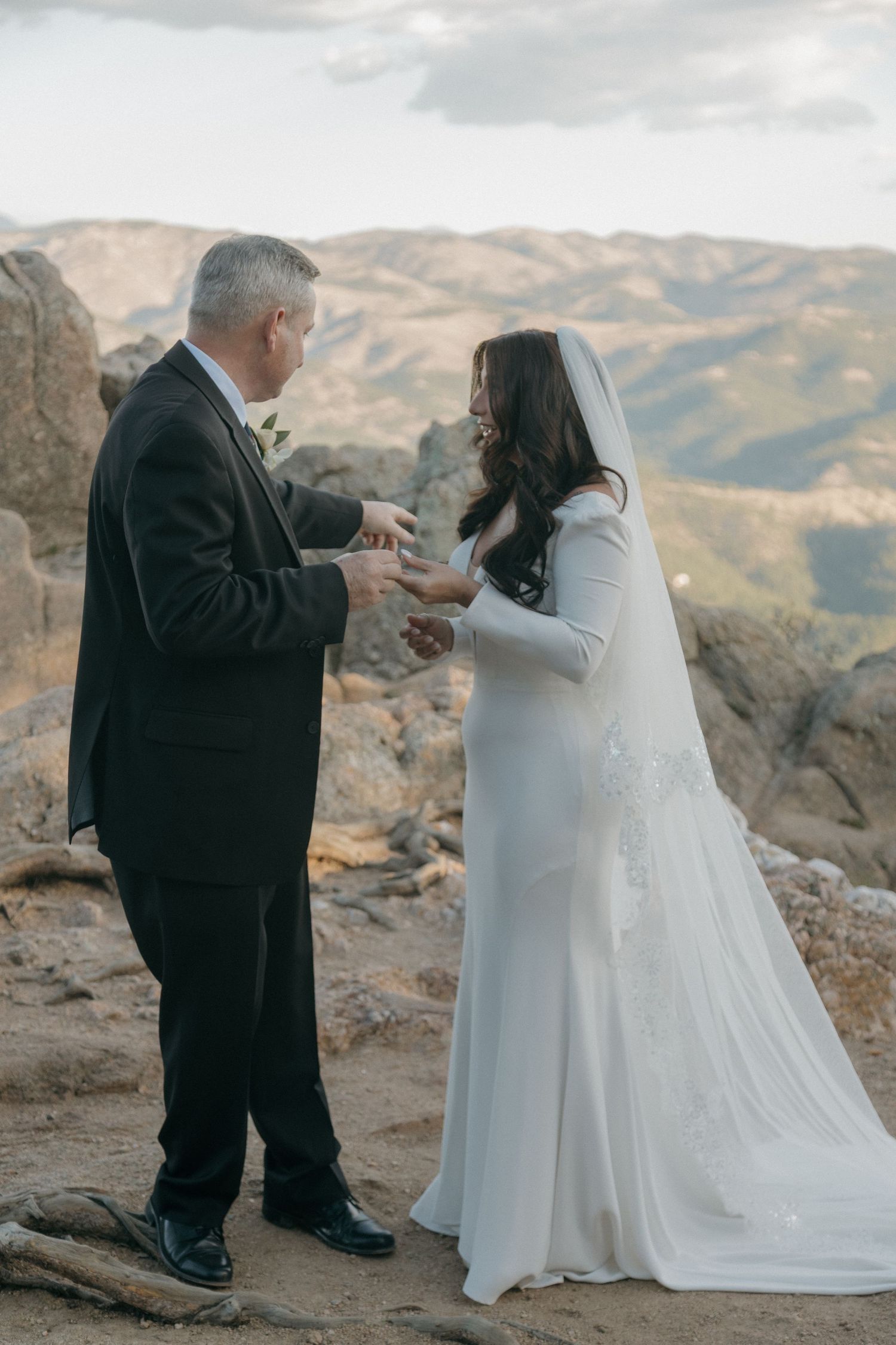 A groom places a wedding ring on the bride's finger during an outdoor mountain ceremony.