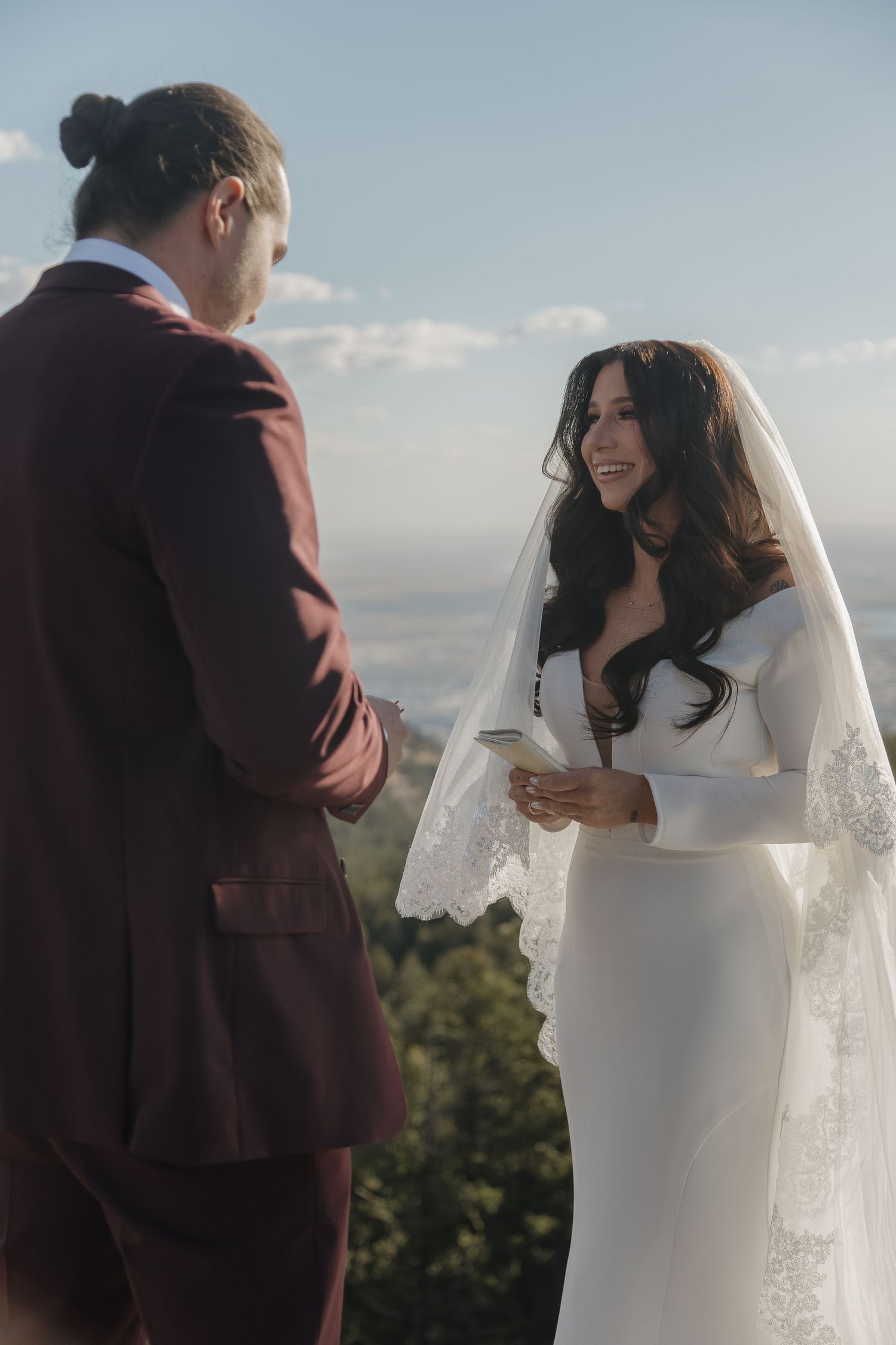 Bride in white long-sleeve gown reading vows to groom in burgundy suit at outdoor mountain elopement.