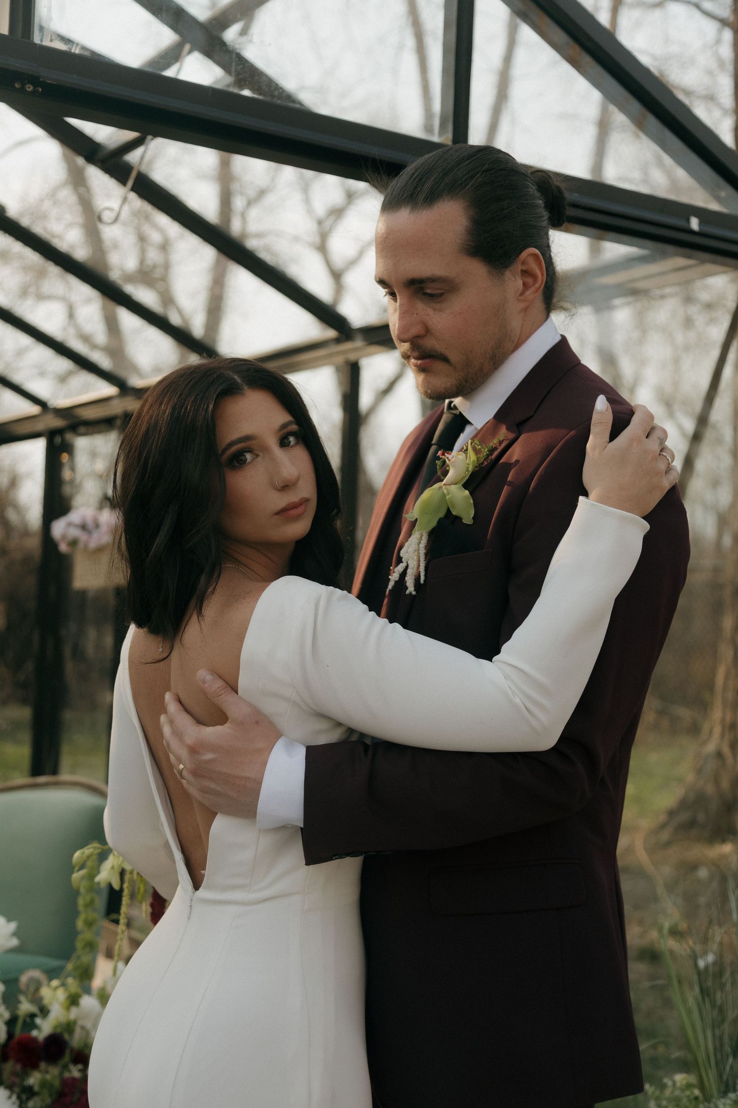 Bride in backless white gown embraces groom in burgundy suit with boutonniere inside a glass greenhouse wedding setting.