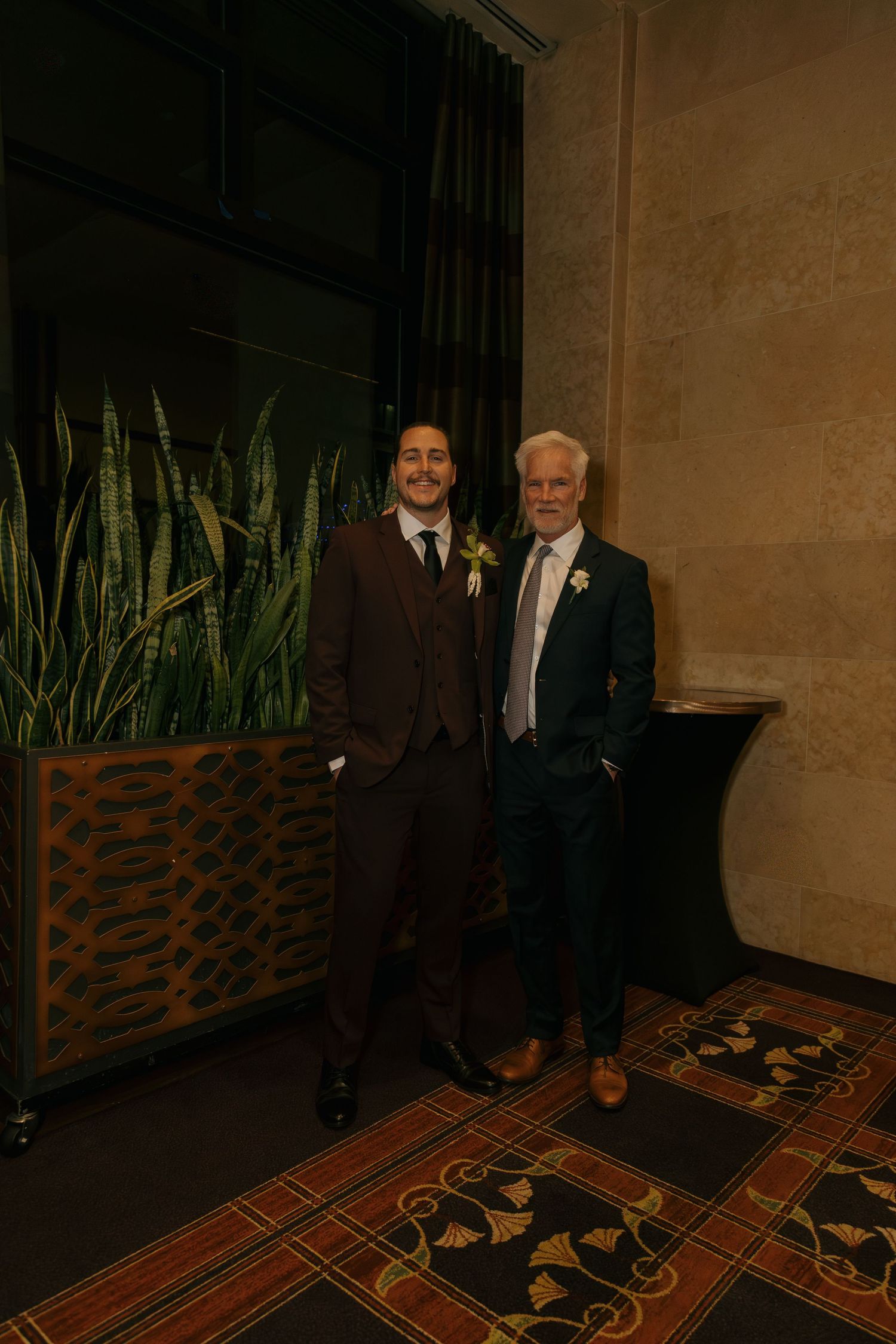 Groom in brown suit posing with older man in teal suit at elegant hotel venue.