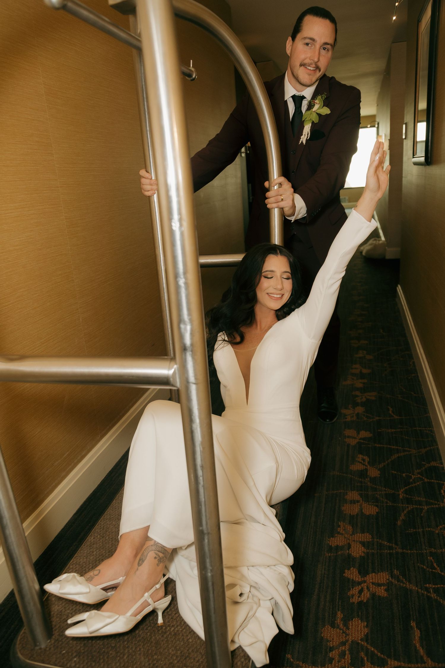Bride in white jumpsuit playfully poses on a luggage cart with groom in hotel hallway.
