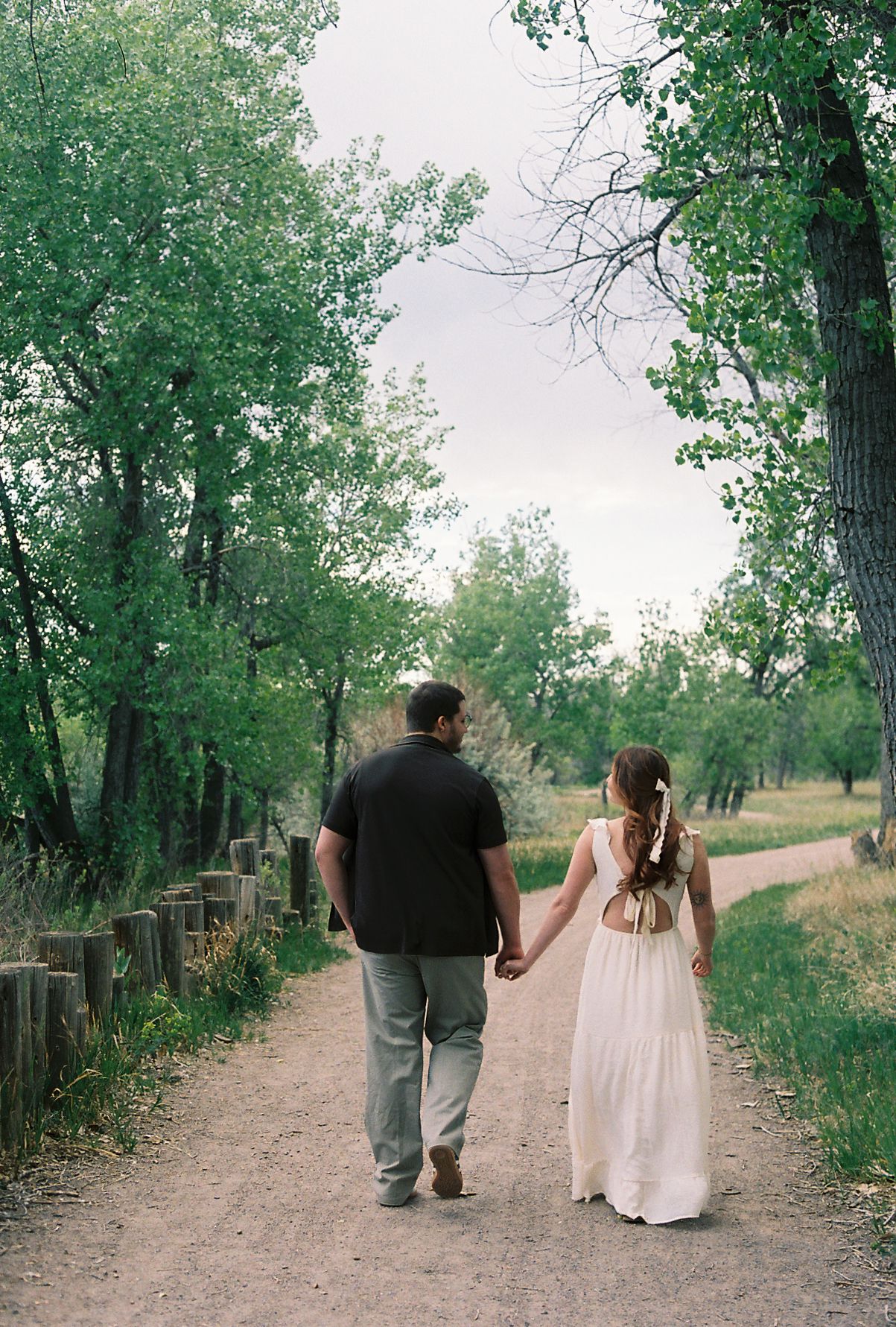 Couple walking hand in hand down a tree-lined dirt path during an outdoor engagement photo session.