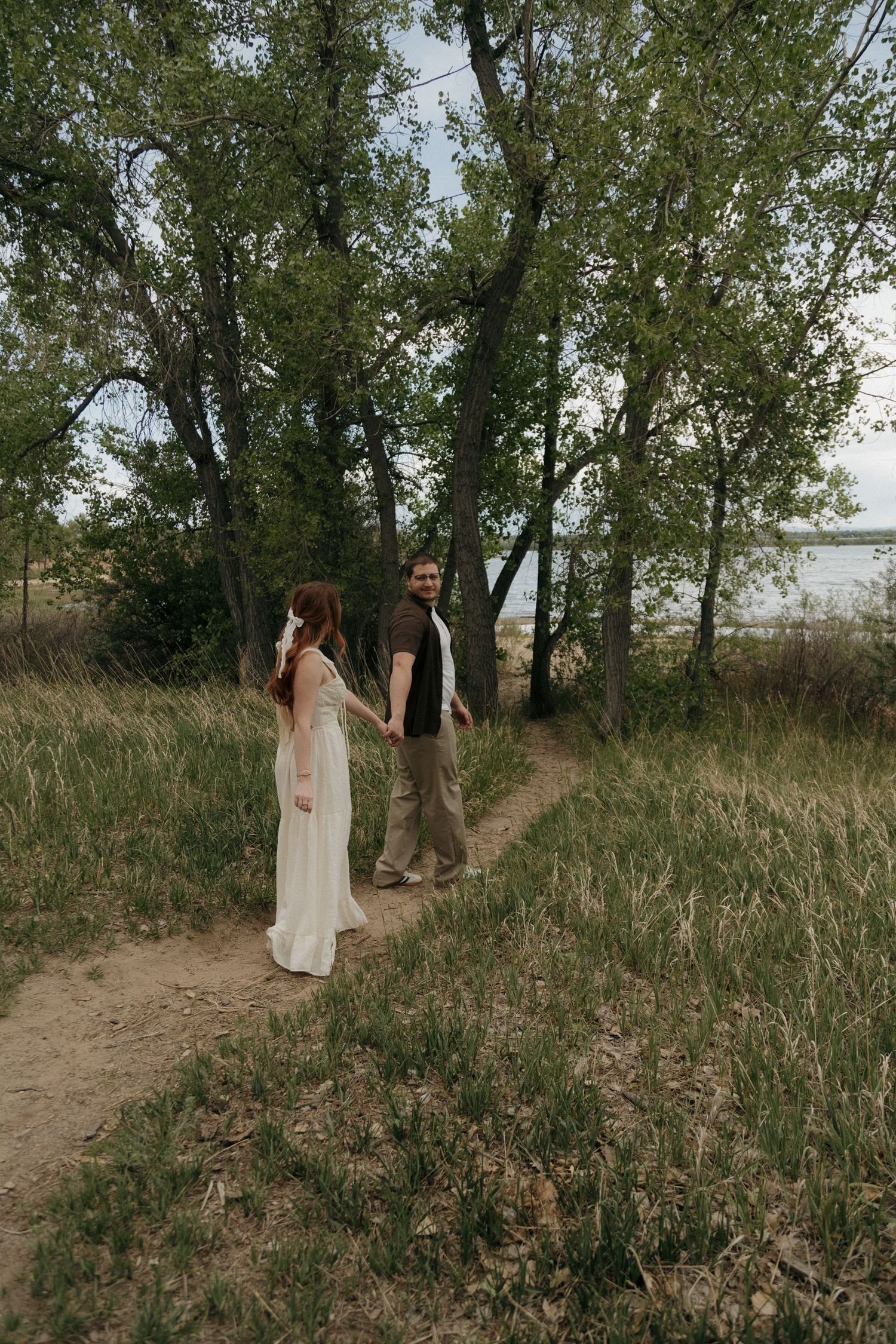 Couple holding hands on a rustic dirt path surrounded by lush green trees during an outdoor engagement session.