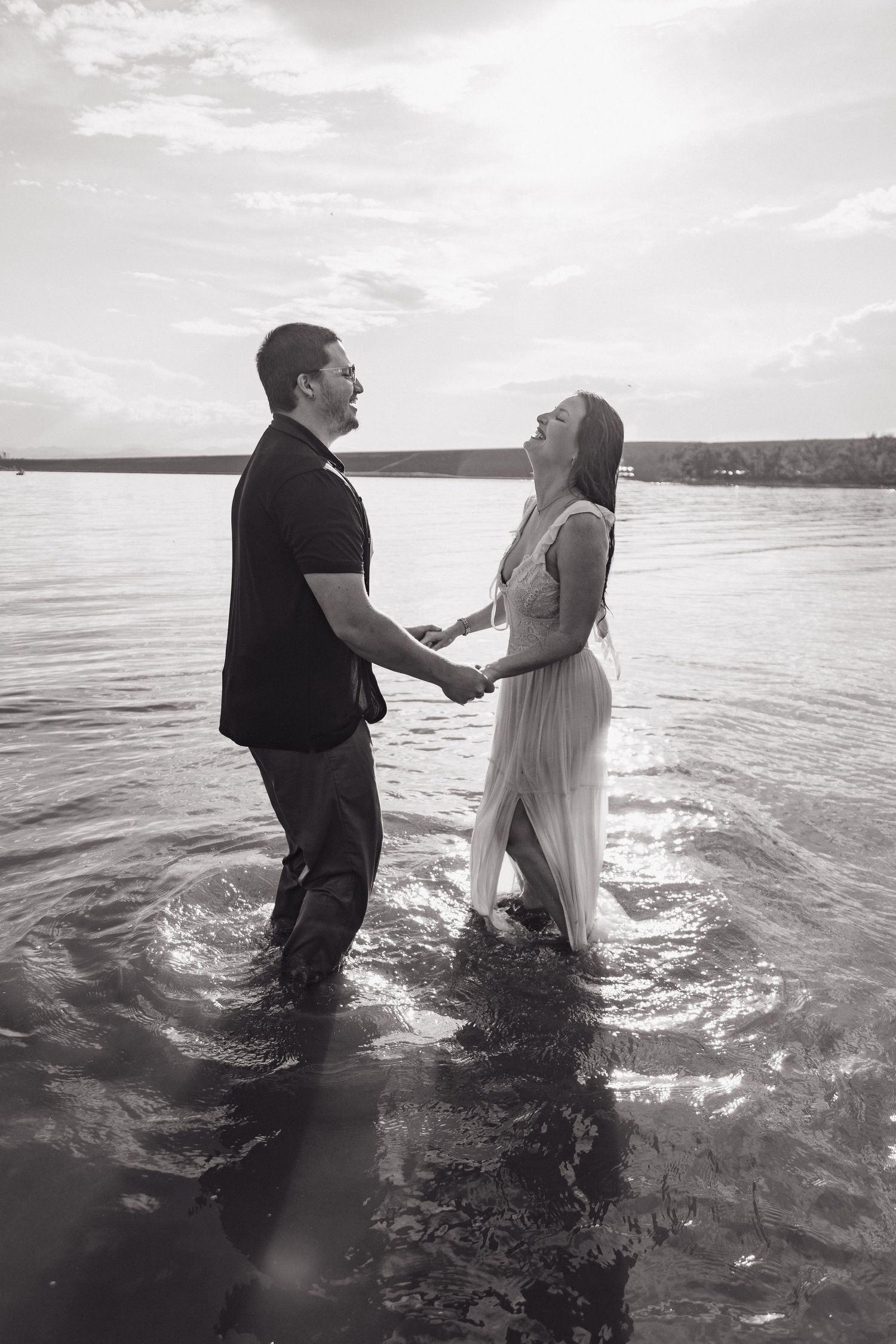 Black and white romantic photo of a couple holding hands and smiling at each other while standing in a calm lake.