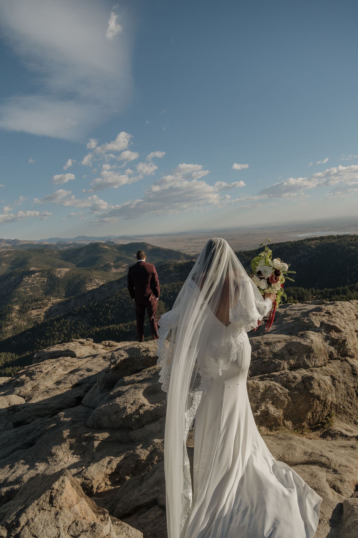 Bride in white gown and veil holds bouquet on rocky mountain summit with groom overlooking vast landscape.