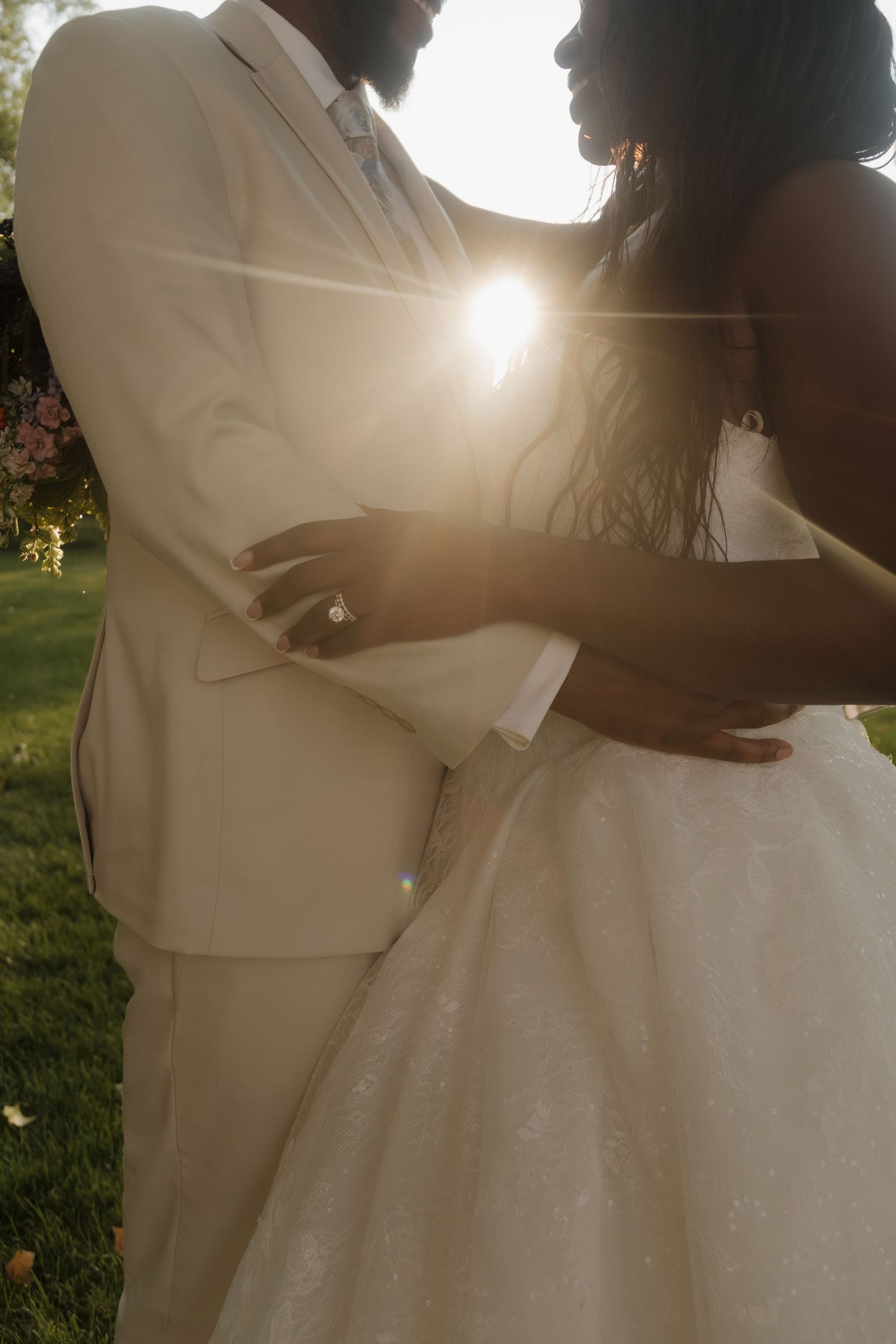 A Black bride and groom embrace in golden sunlight, wearing white wedding attire outdoors.
