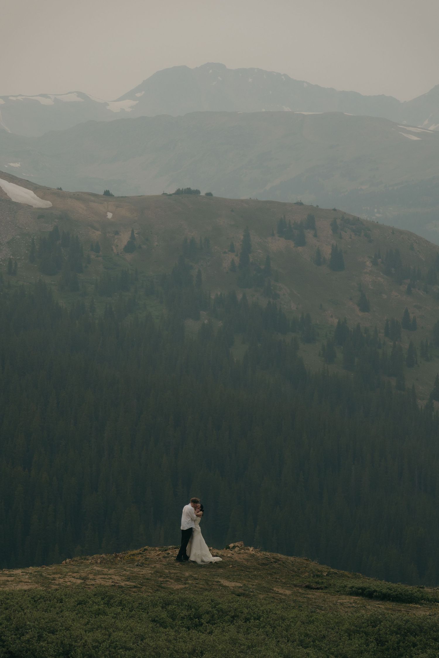 Bride and groom embrace on a mountain summit with sweeping layered forest and misty mountain views behind them.