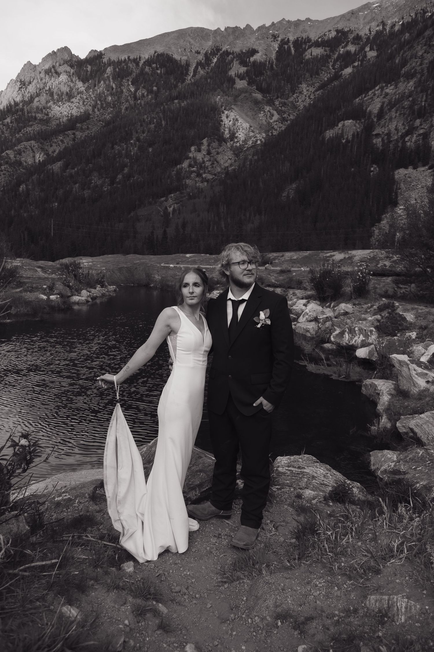 Black and white portrait of bride and groom standing by a mountain lake after wedding.