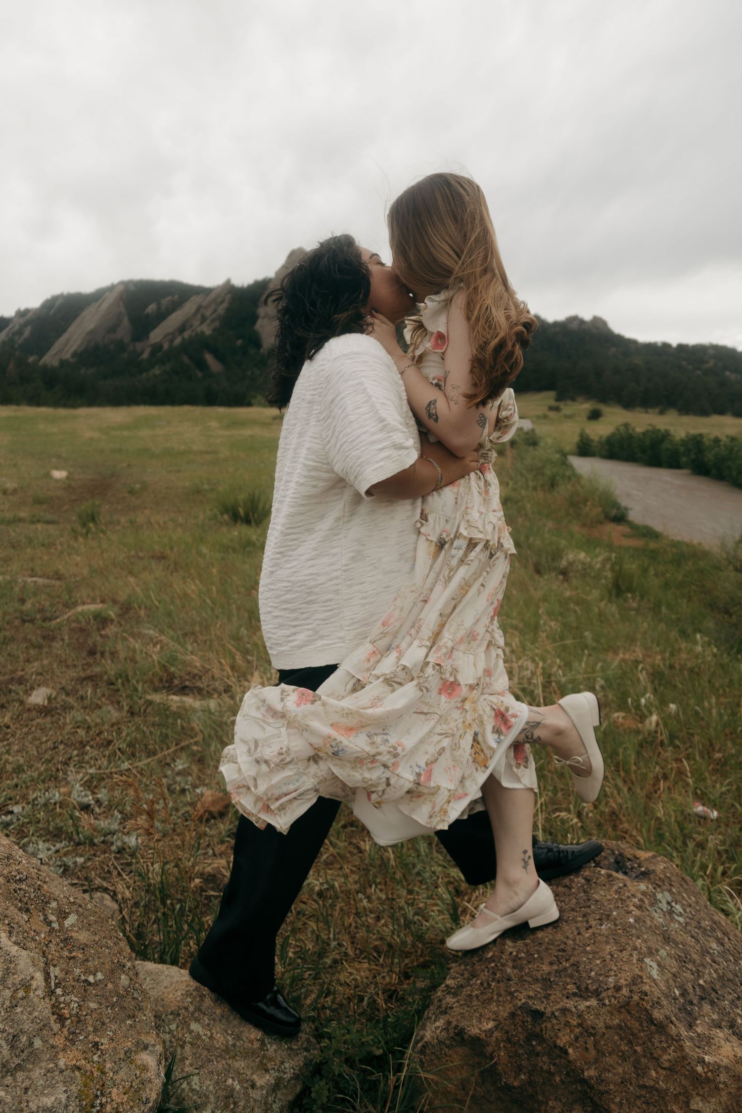 Two women share a passionate kiss while standing on rocks in a scenic mountain meadow landscape.