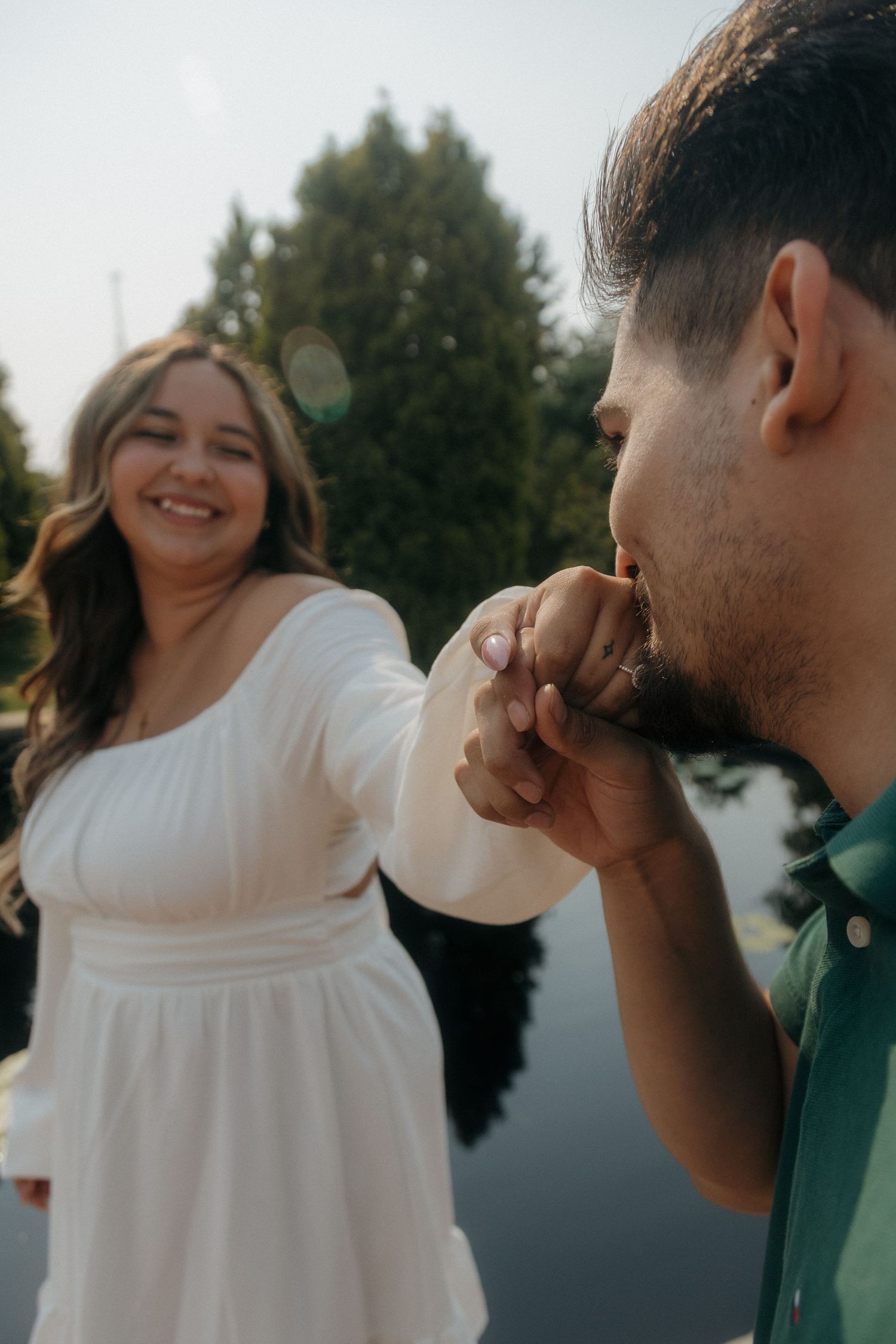 A man kisses a smiling woman's hand outdoors, she wears a white dress near a reflective pond with trees.