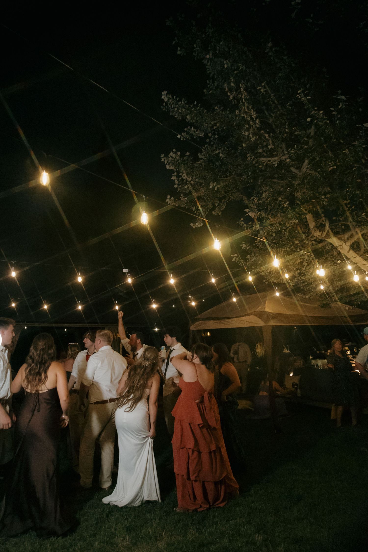 Outdoor wedding reception at night under a clear tent with string lights, guests dancing on grass surrounded by trees.