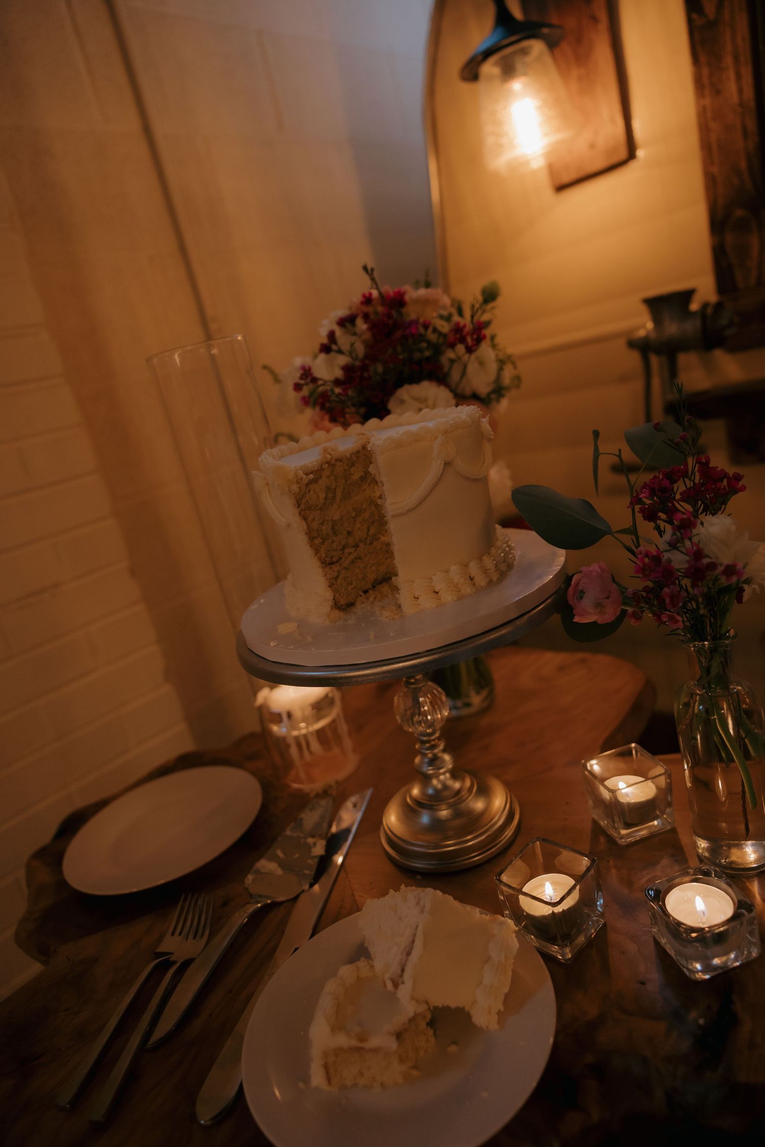 Single-tier white wedding cake with floral topper surrounded by candles on rustic wooden table.