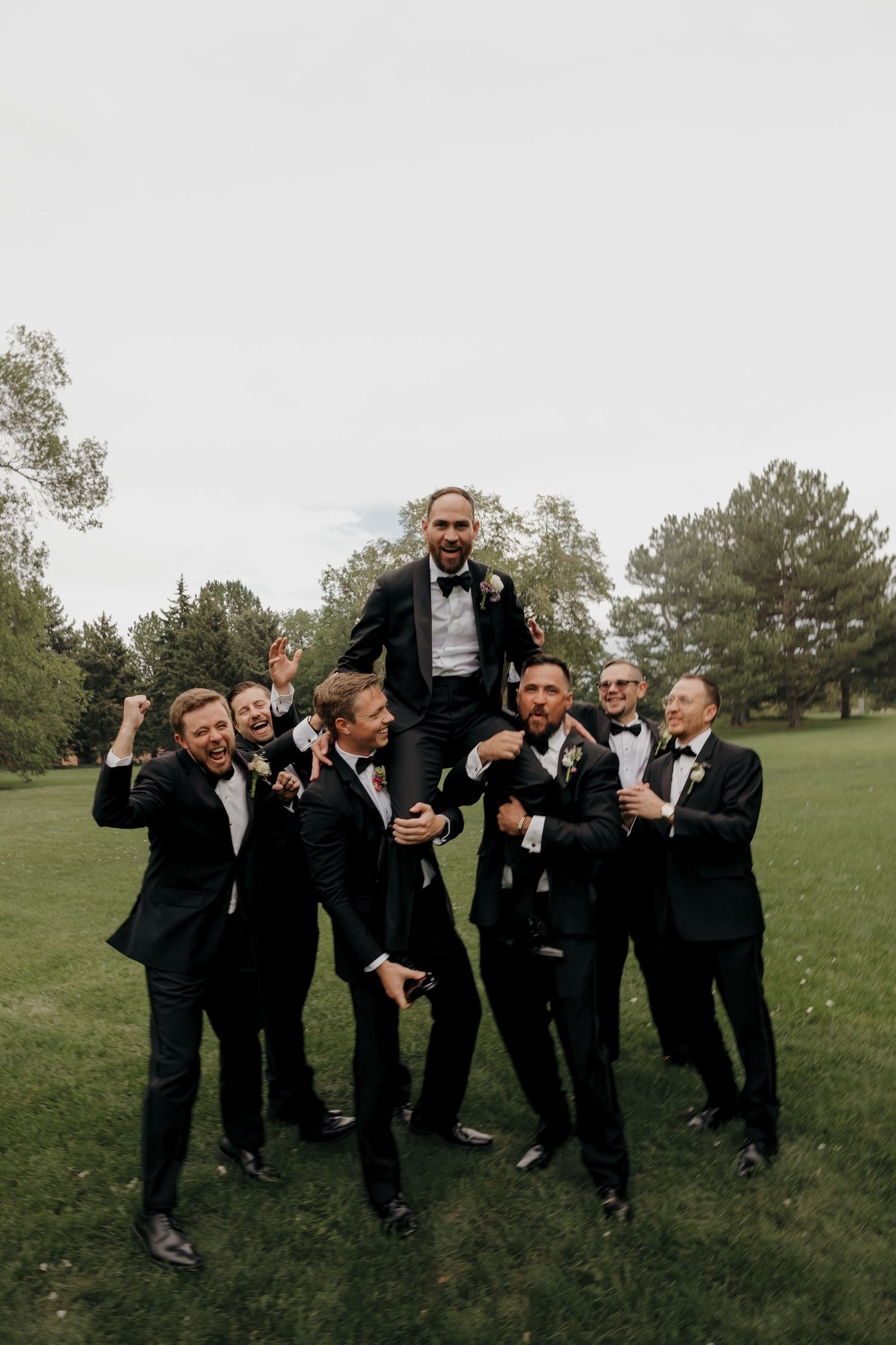 Groom lifted on shoulders by groomsmen in black tuxedos celebrating outdoors on a green lawn.
