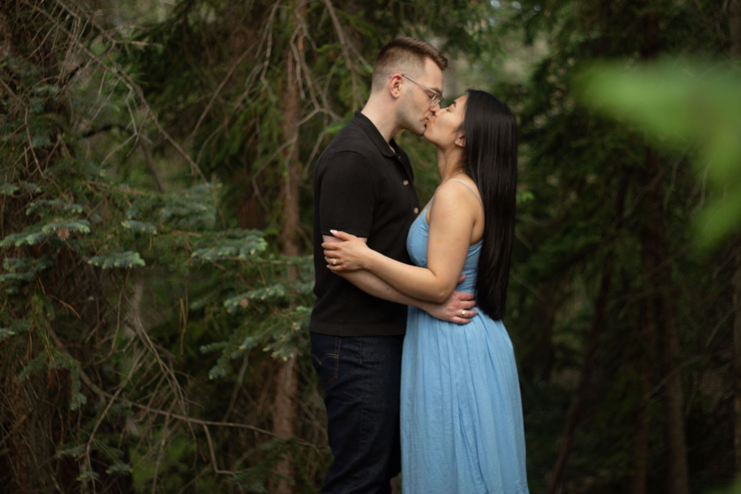 Two people share an intimate embrace against a backdrop of evergreen trees.