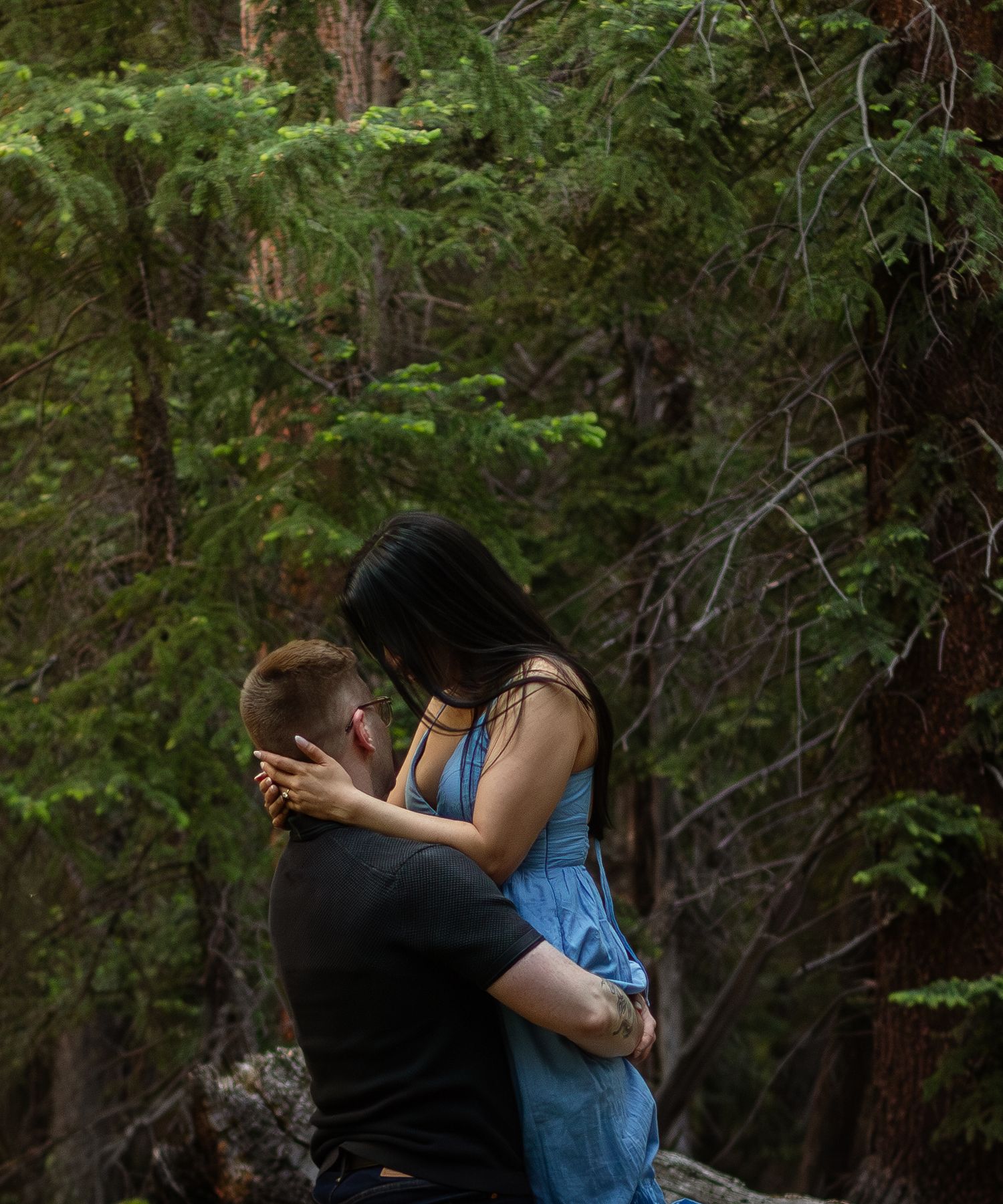 A couple shares a romantic kiss in a forest setting with dramatic lighting and pine trees.
