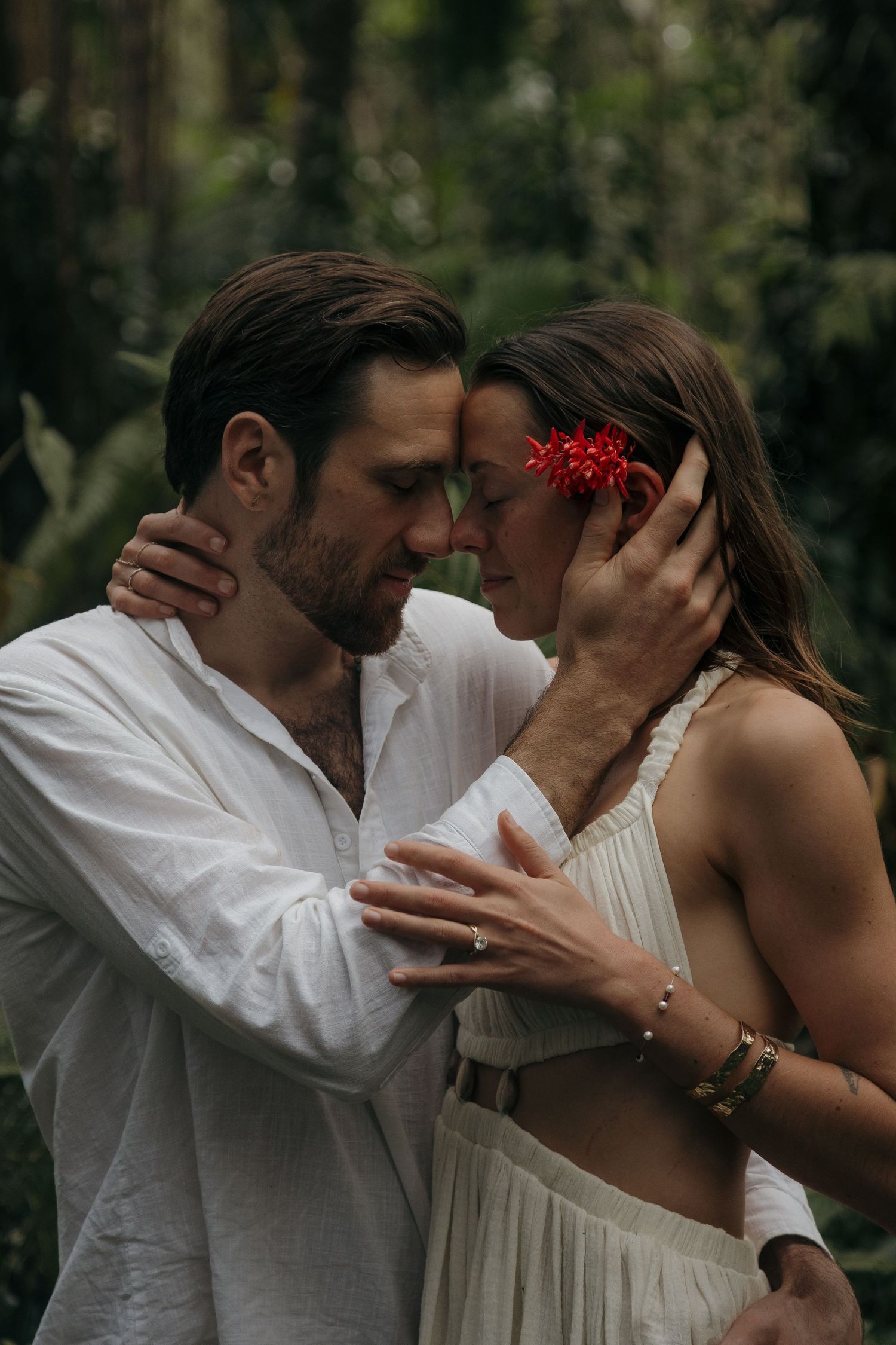 Romantic couple in white clothing share an intimate moment in a lush garden setting with a red flower accent.