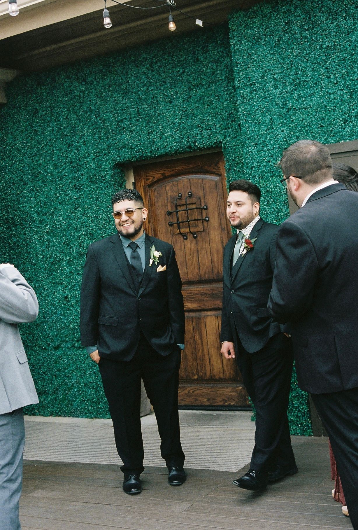 Two groomsmen in black suits with boutonnieres smiling and conversing in front of green hedge wall backdrop.