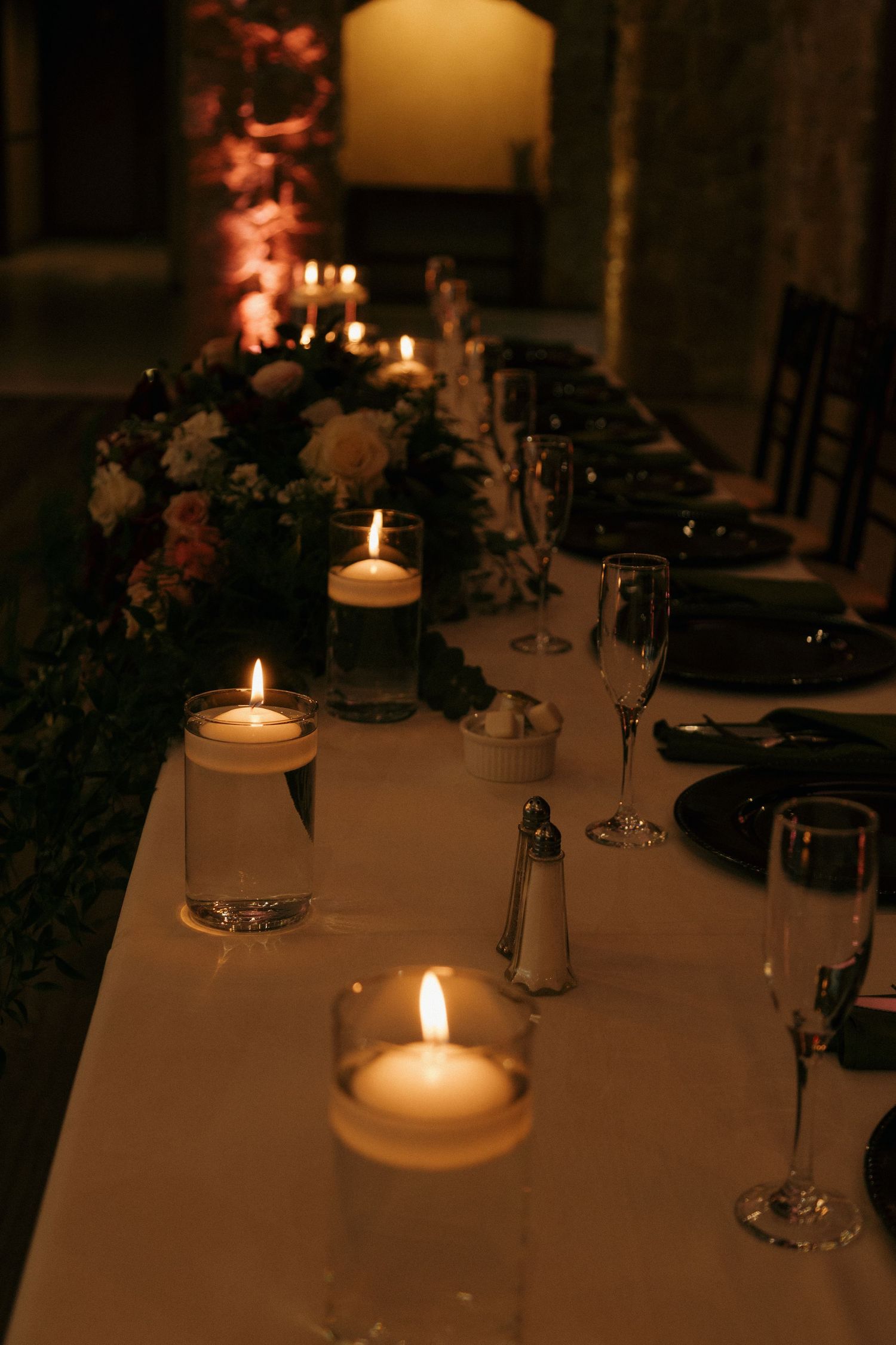 Long wedding reception table with floating candles, floral garland, black plates in dimly lit stone hall.