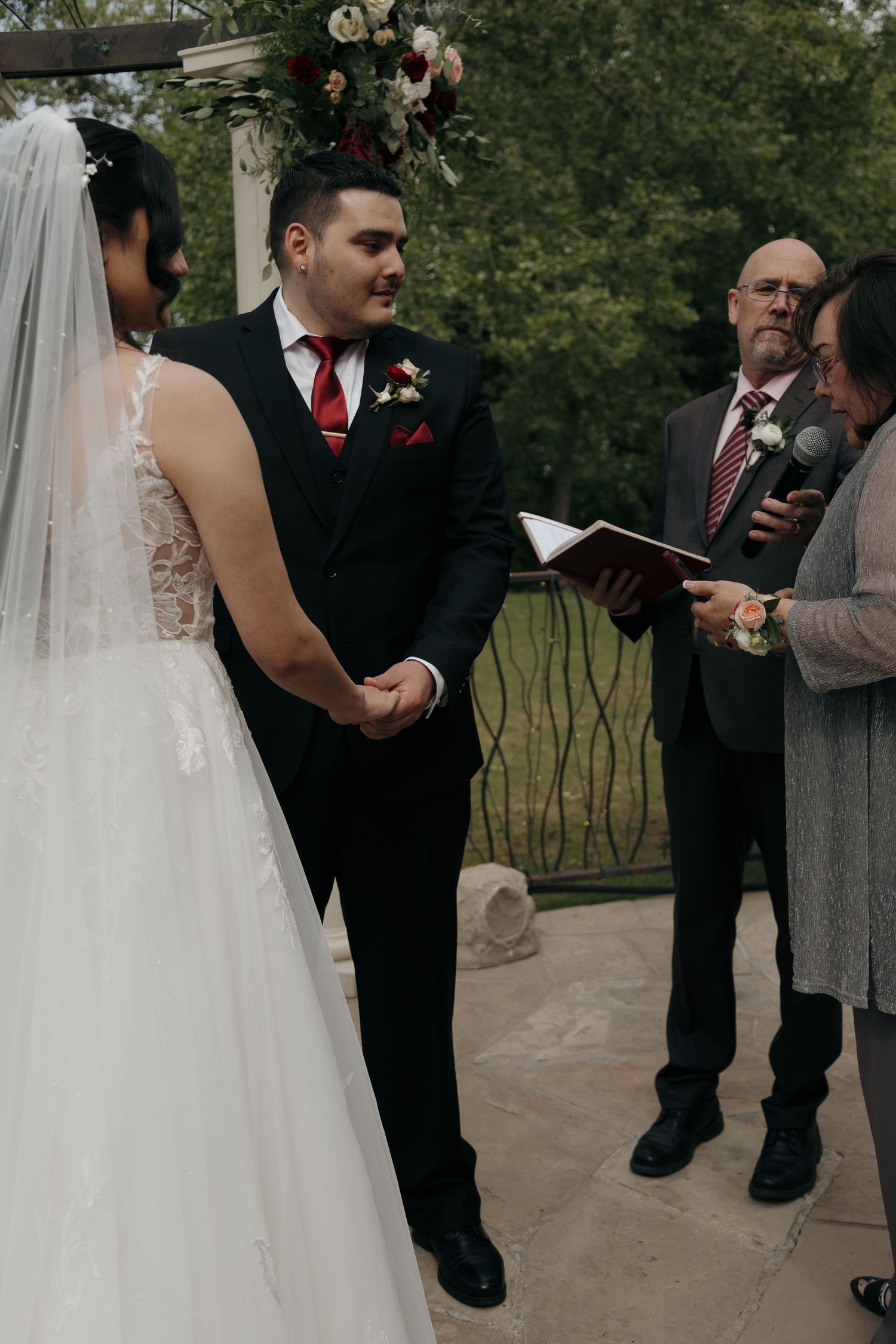 Groom in black suit with red tie holds brides hands during outdoor wedding ceremony with officiant.