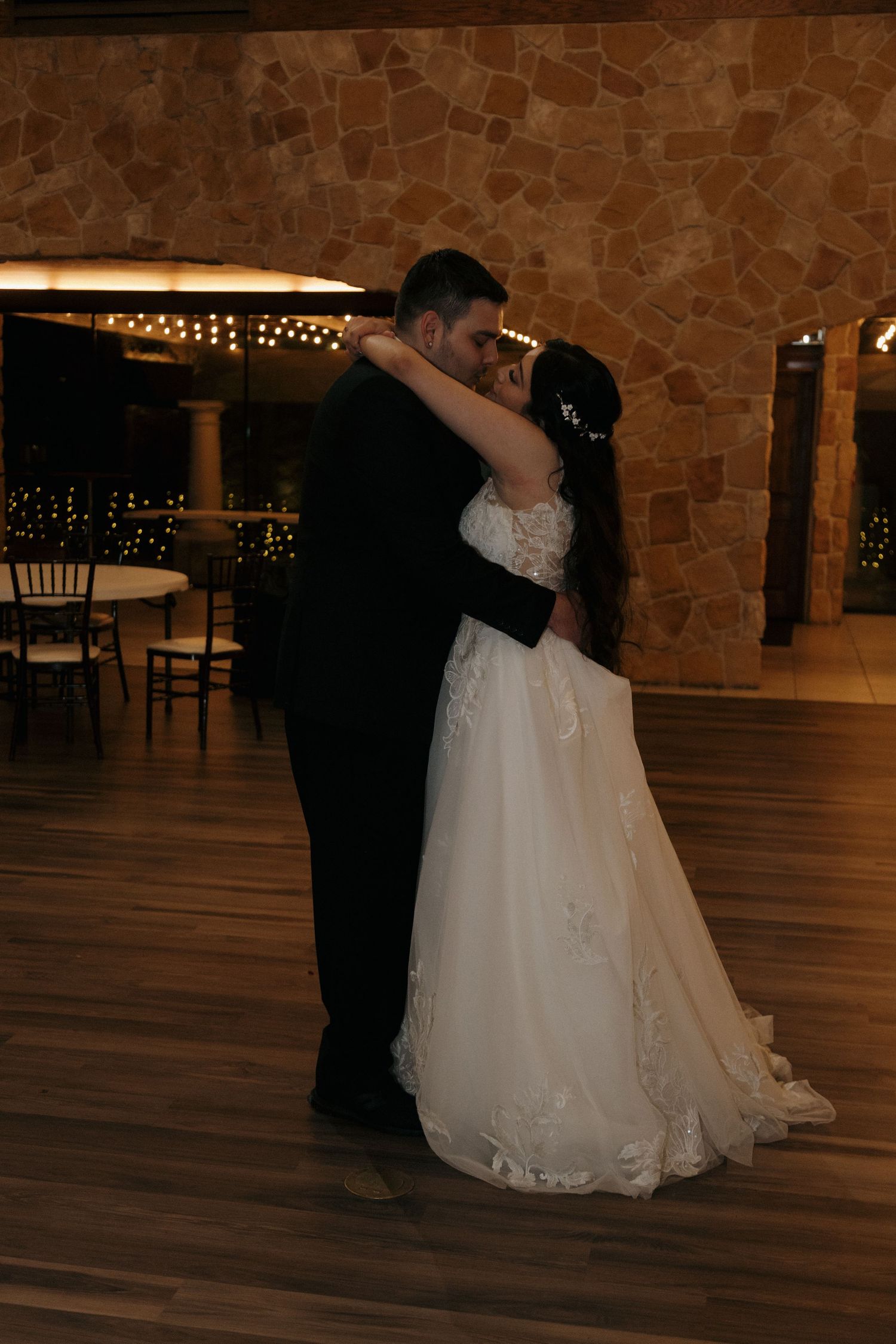 A bride and groom share a romantic slow dance embrace in a warmly lit rustic stone wedding venue.