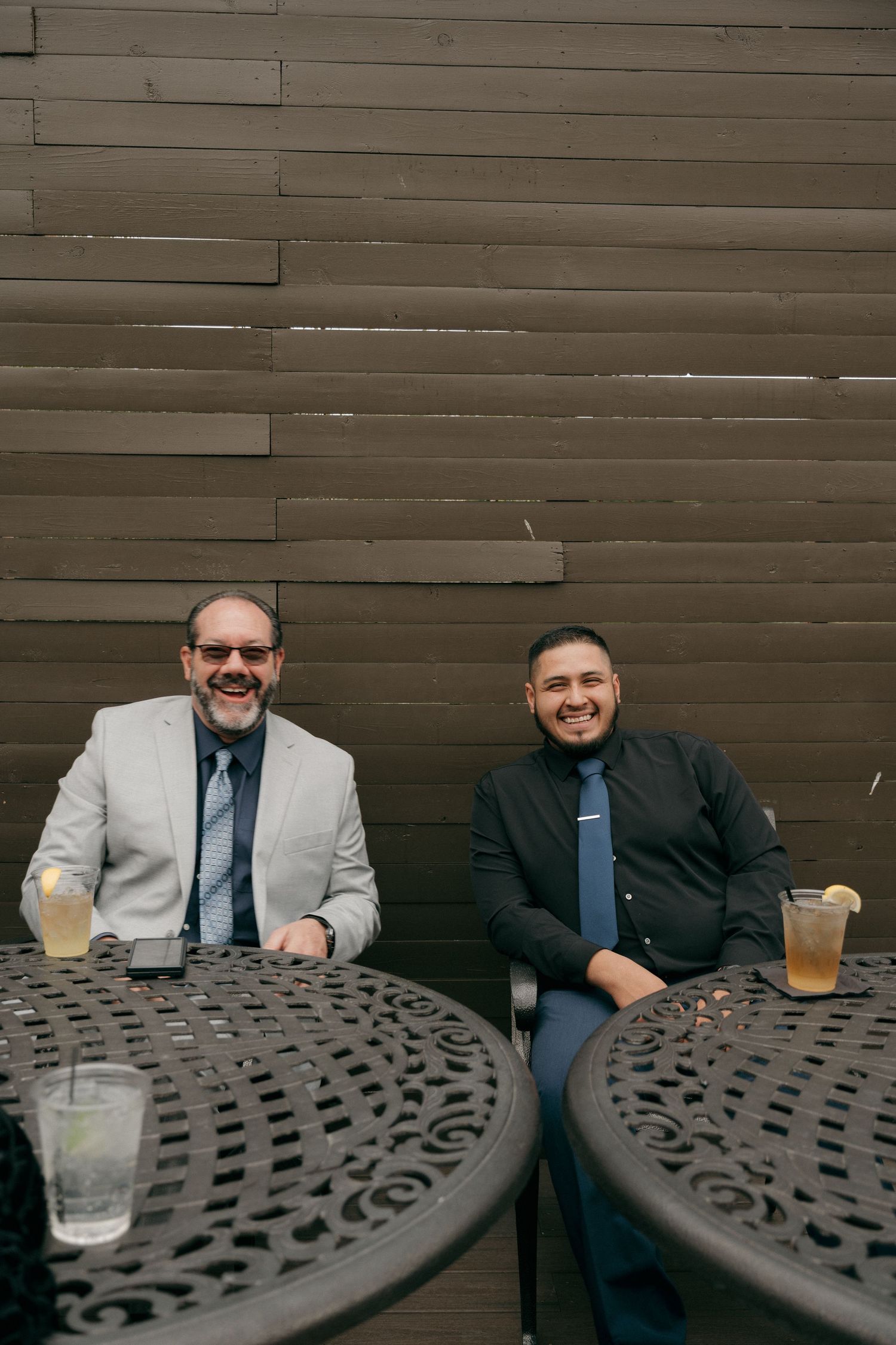 Two men in suits smile while seated at an ornate outdoor table with cocktails at a wedding reception.