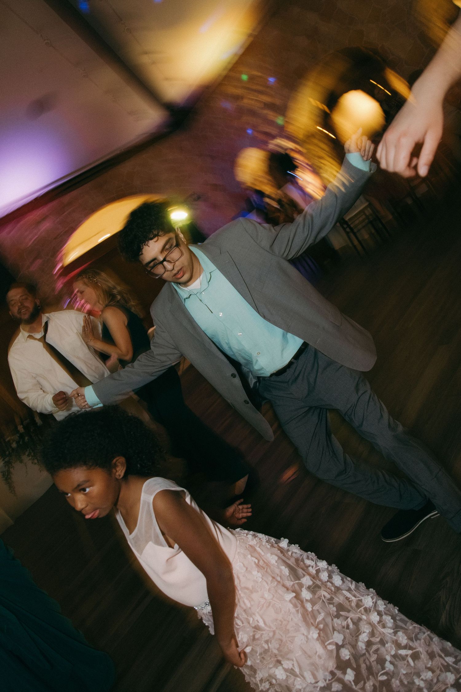Wedding guest in gray suit dances energetically on a colorfully lit reception dance floor.