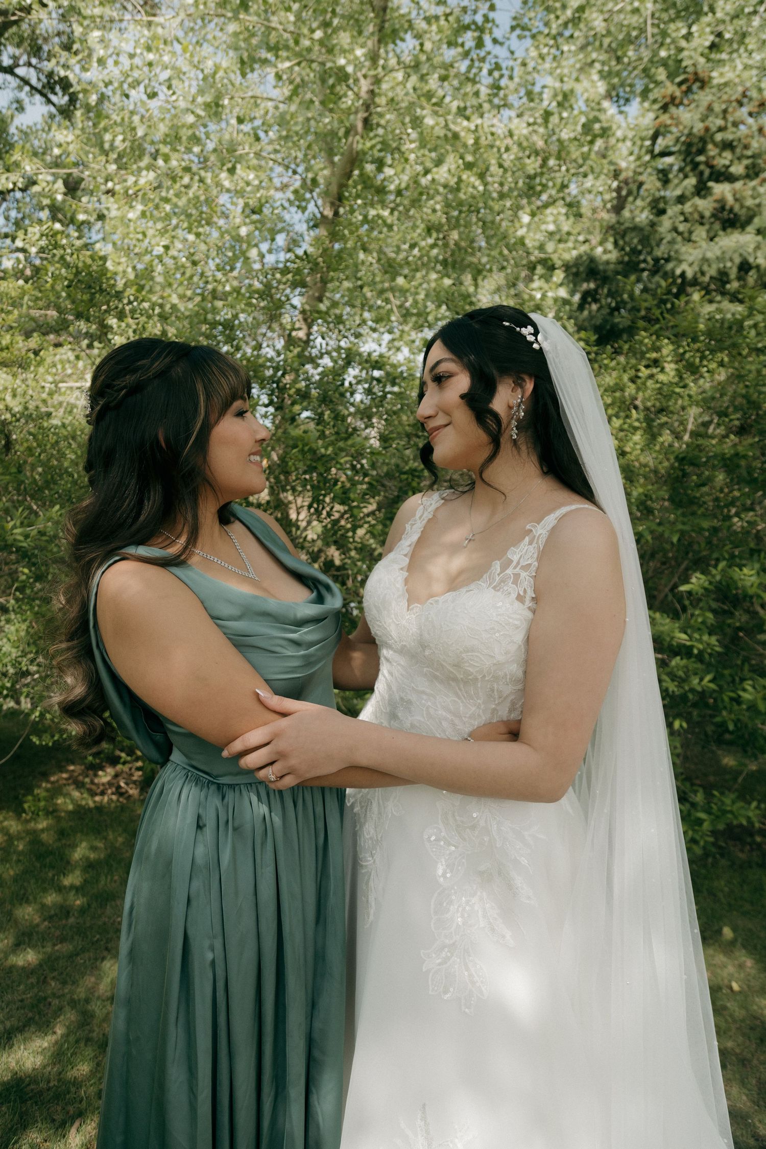 Bride in lace gown and bridesmaid in sage green dress share a loving gaze surrounded by lush greenery.