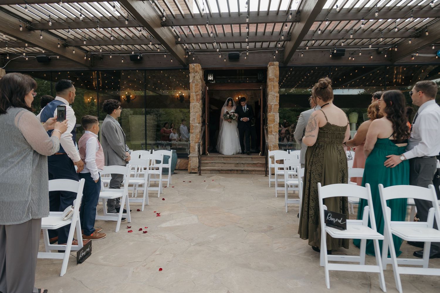 Wedding guests watching bride and groom at altar during outdoor ceremony under wooden pergola with white chairs.