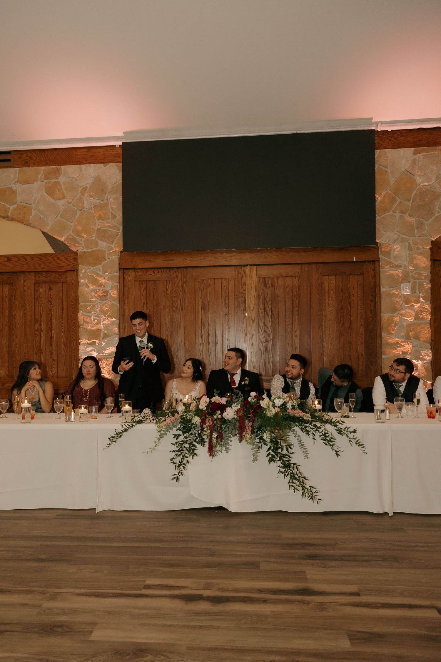 Wedding party seated at decorated head table during indoor reception with stone wall backdrop.