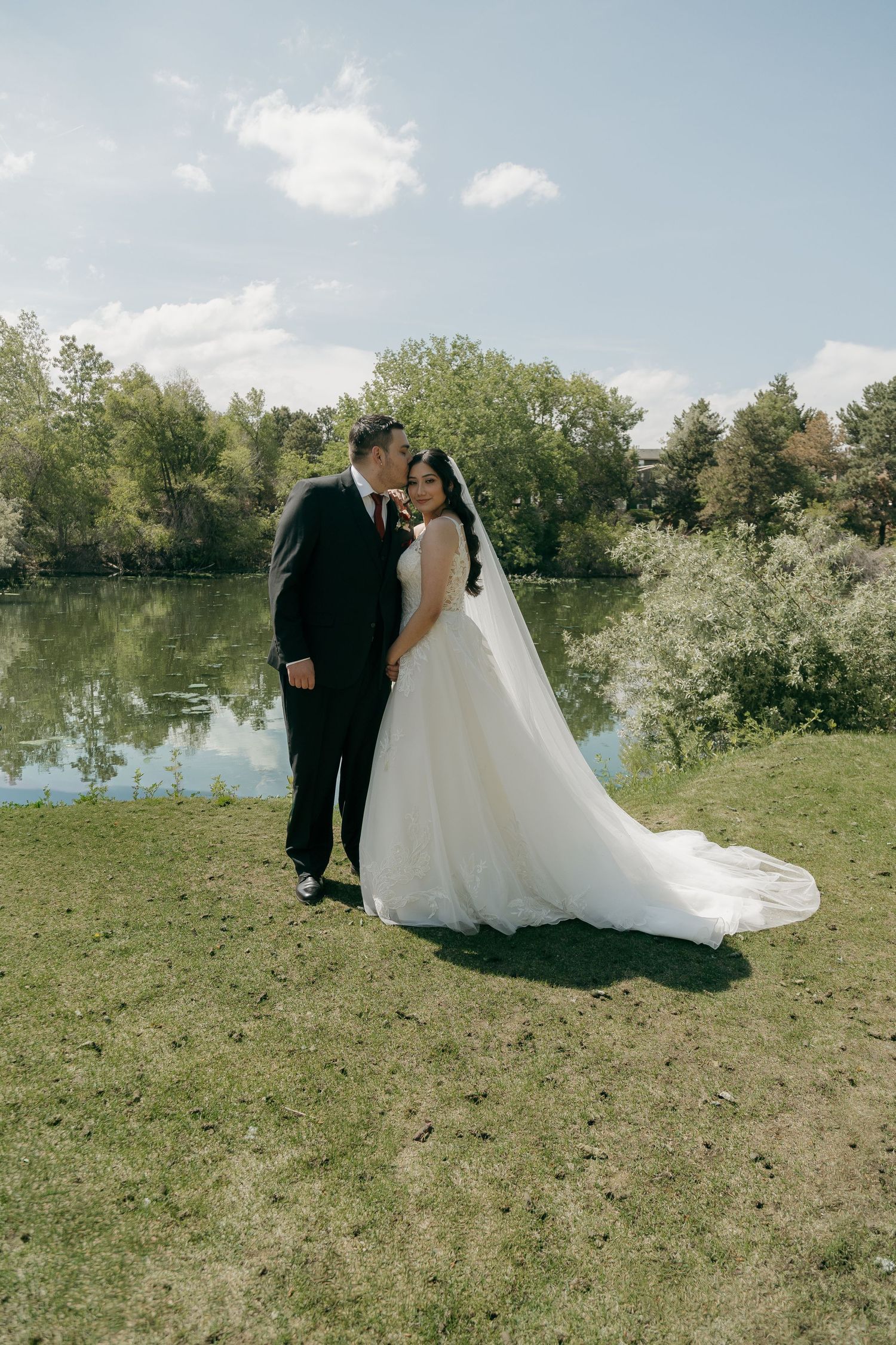 Bride and groom posing by a serene lake, groom kissing bride's cheek on sunny wedding day.
