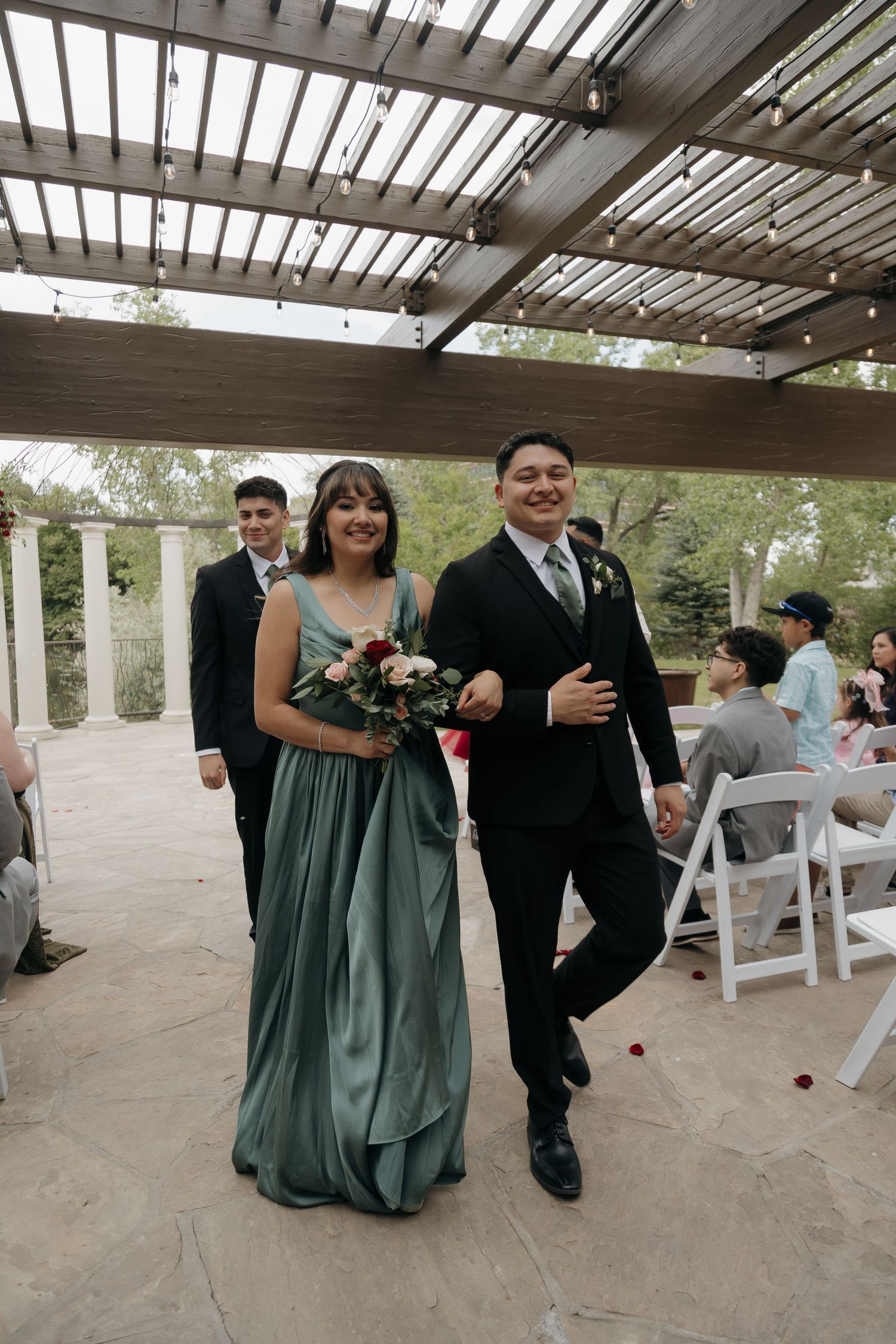 Bridesmaid in sage green dress walks with groomsman in black suit down wedding aisle.