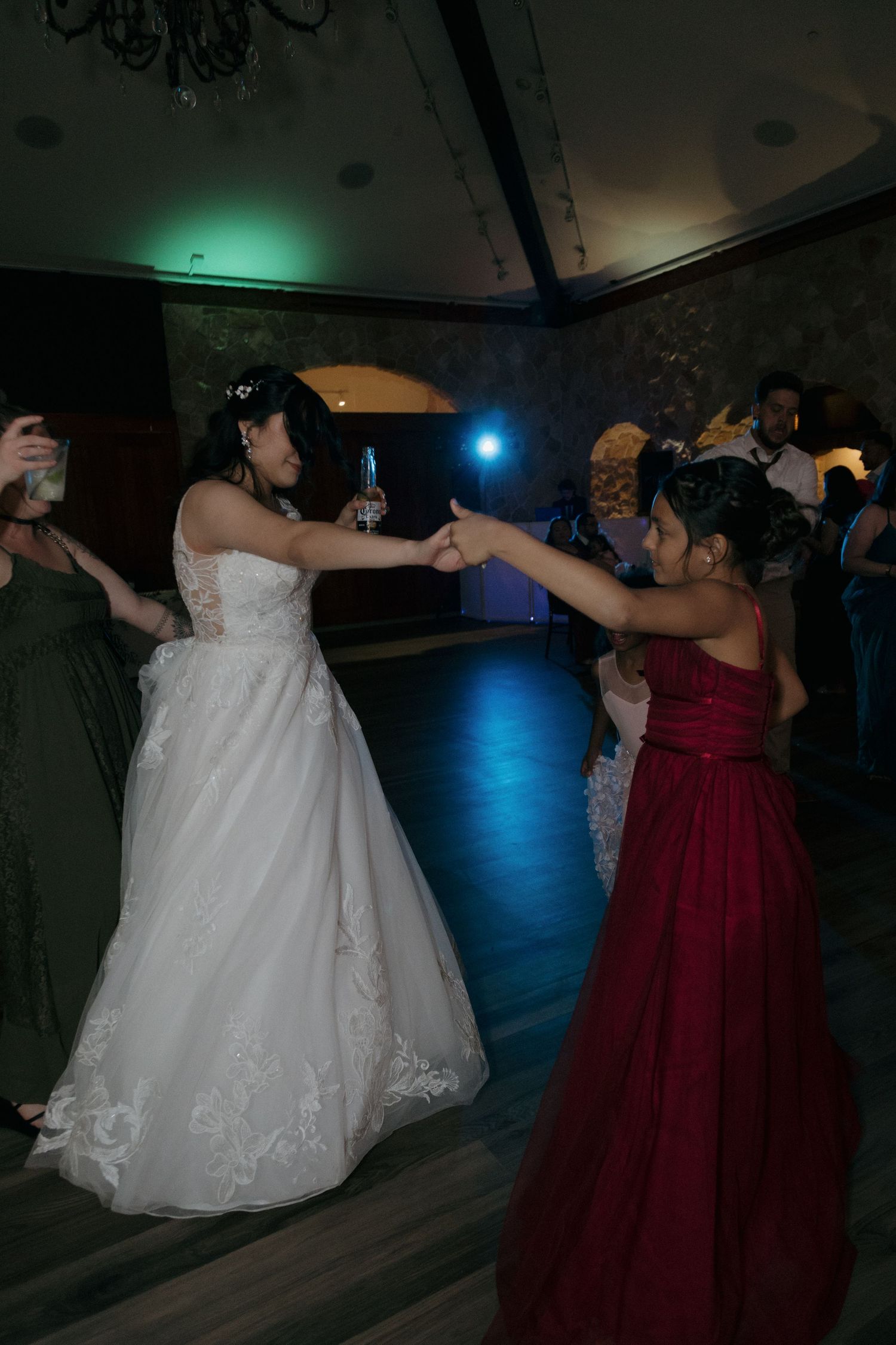 Bride in white strapless gown dances with woman in red dress on colorfully lit wedding reception floor.
