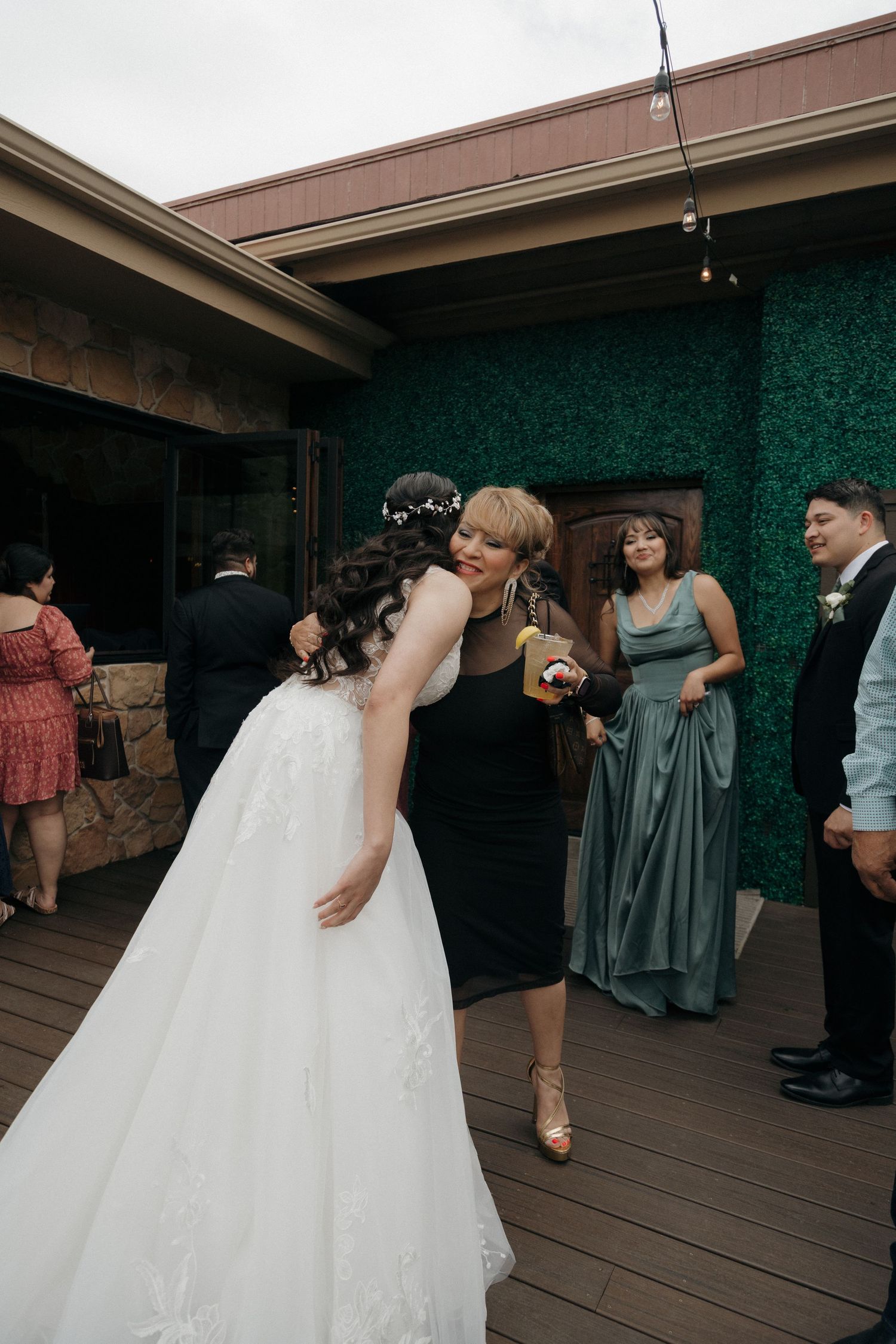 Bride in white lace gown embracing guest in black dress at outdoor wedding reception.