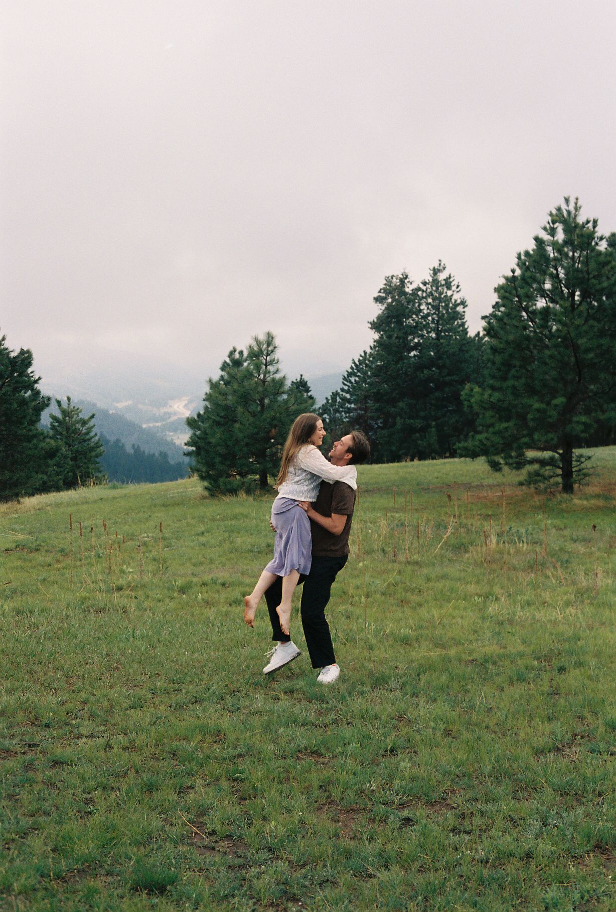 A couple embraces in a lush green meadow surrounded by pine trees and misty mountains in the background.