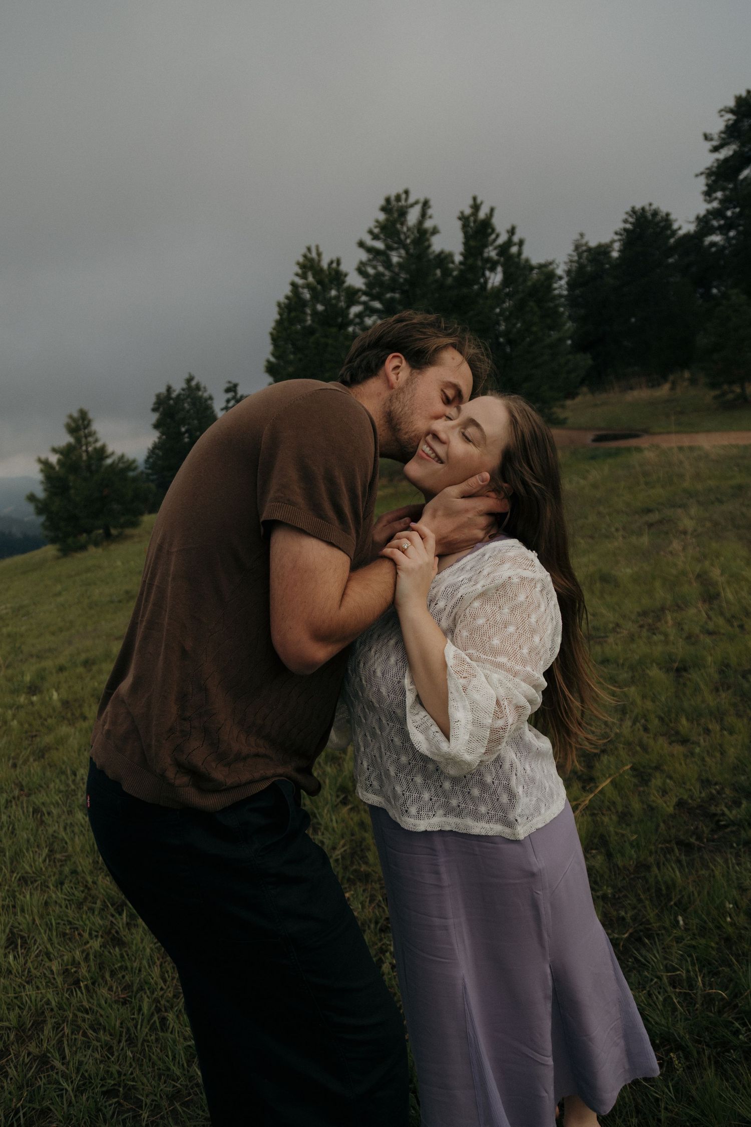 A man kisses a woman on the cheek as they hold hands in a green meadow.