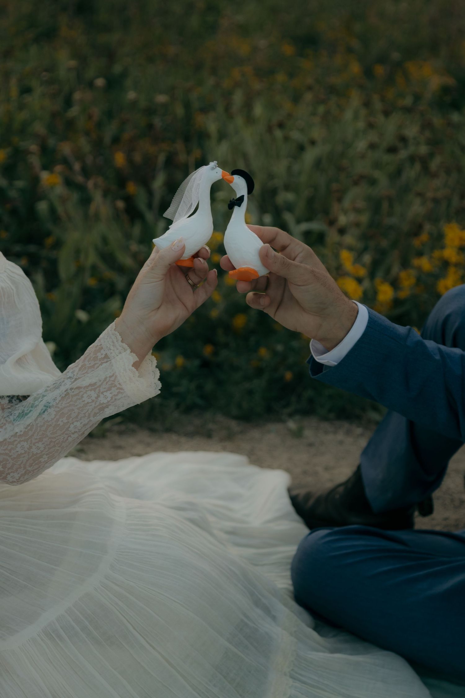 Bride and groom holding decorative swan figurines together in a sunny flower field.