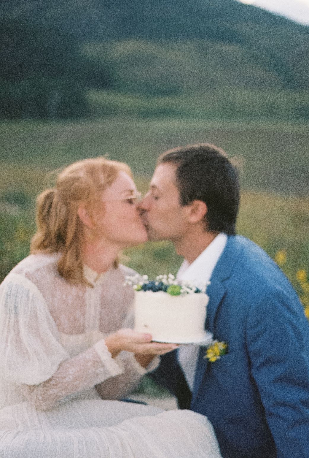 Bride and groom share a kiss while holding a white wedding cake in a sunny mountain meadow.