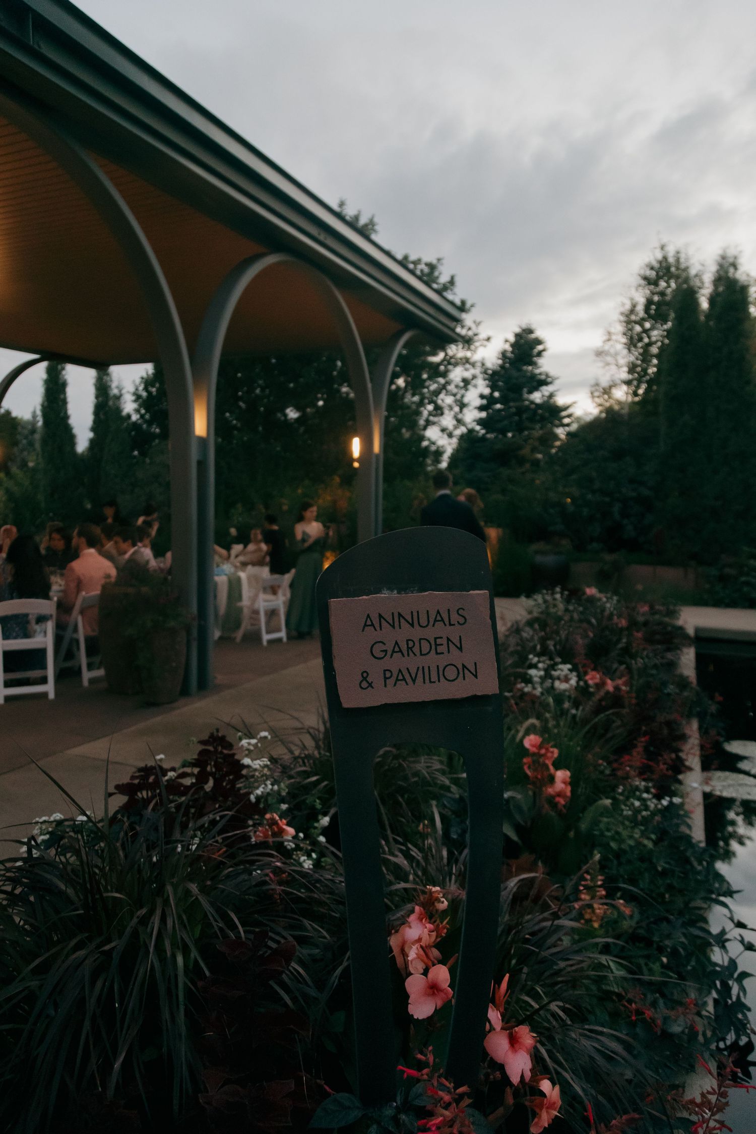 Sign for Annuals Garden and Pavilion with wedding reception guests seated under pavilion at dusk.