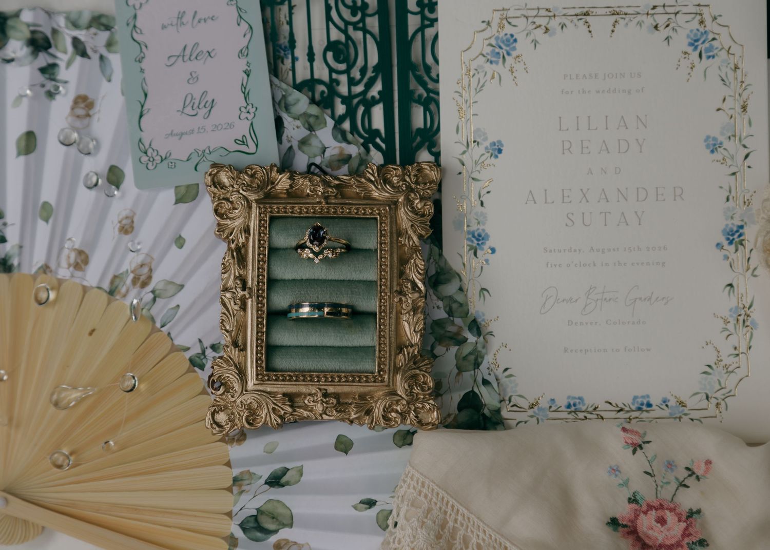 Wedding flat lay featuring gold ornate frame with rings, floral invitation suite for Lilian Brady and Alexander Retay, and decorative fans.