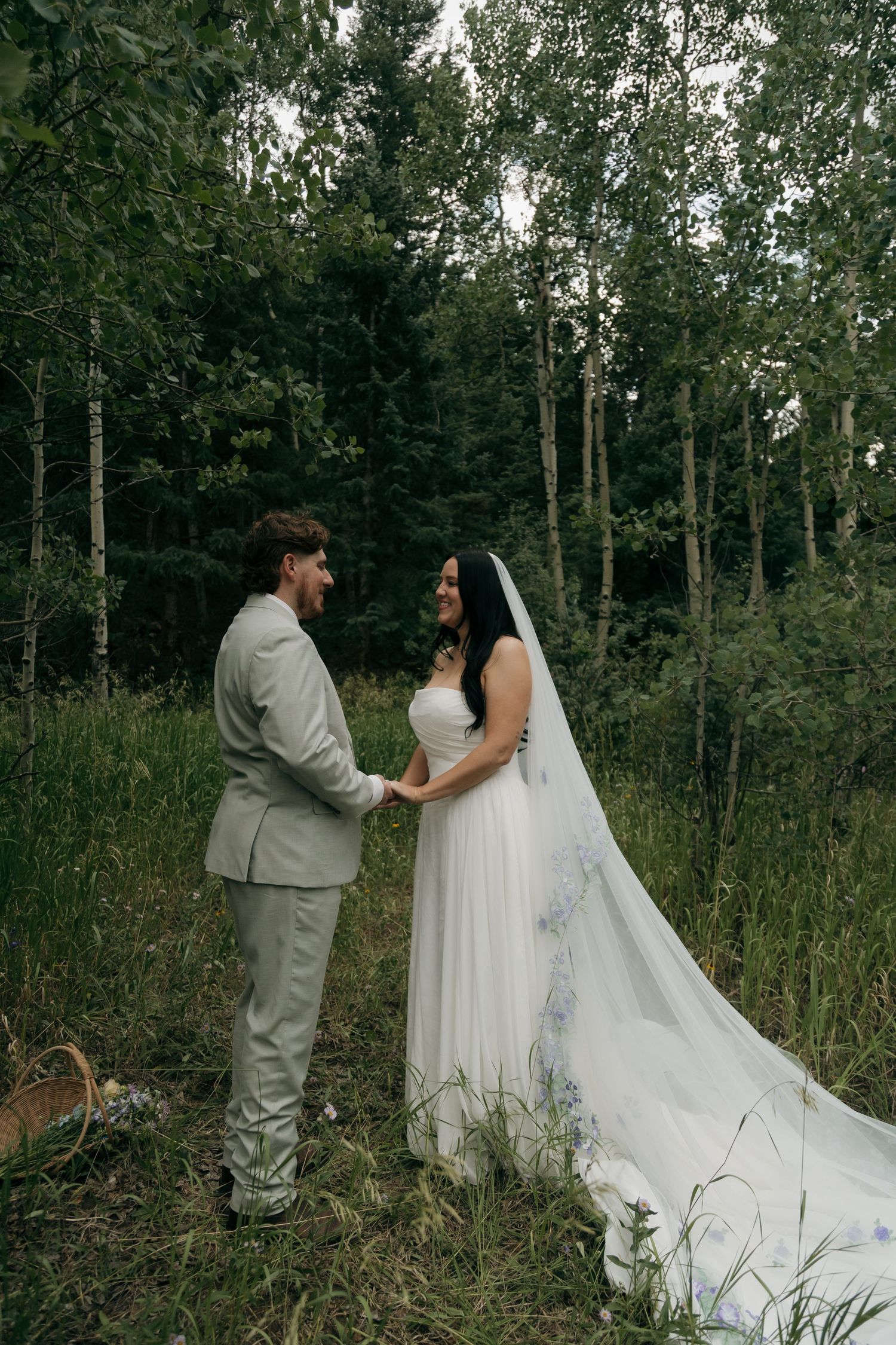 Bride and groom hold hands smiling at each other in a lush forest during their outdoor wedding.