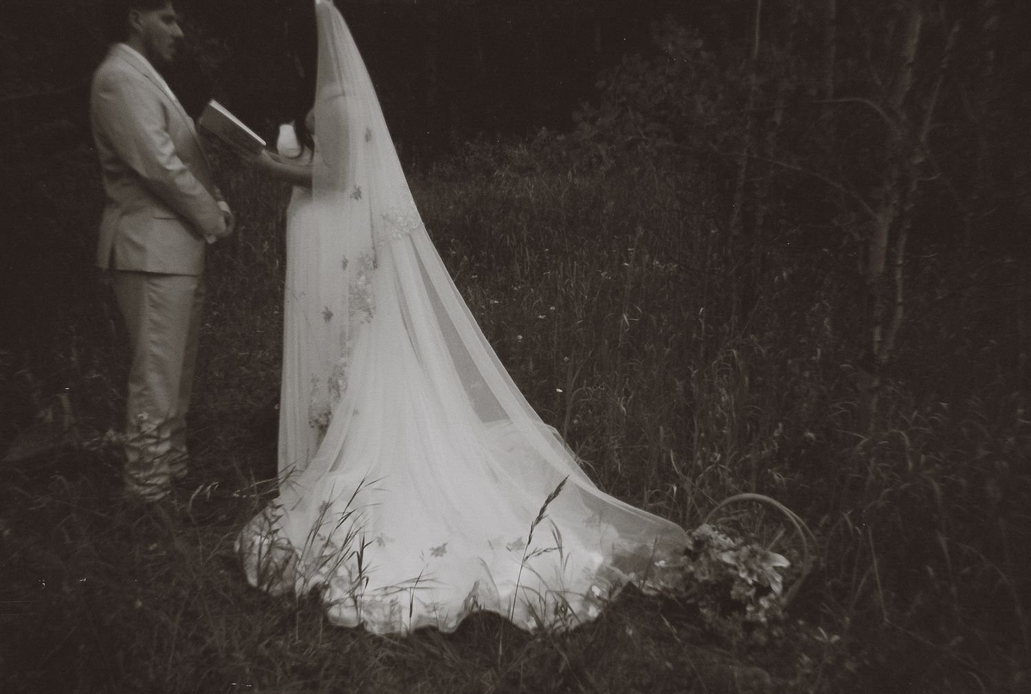 Vintage black and white photo of bride in flowing veil during outdoor forest wedding ceremony.