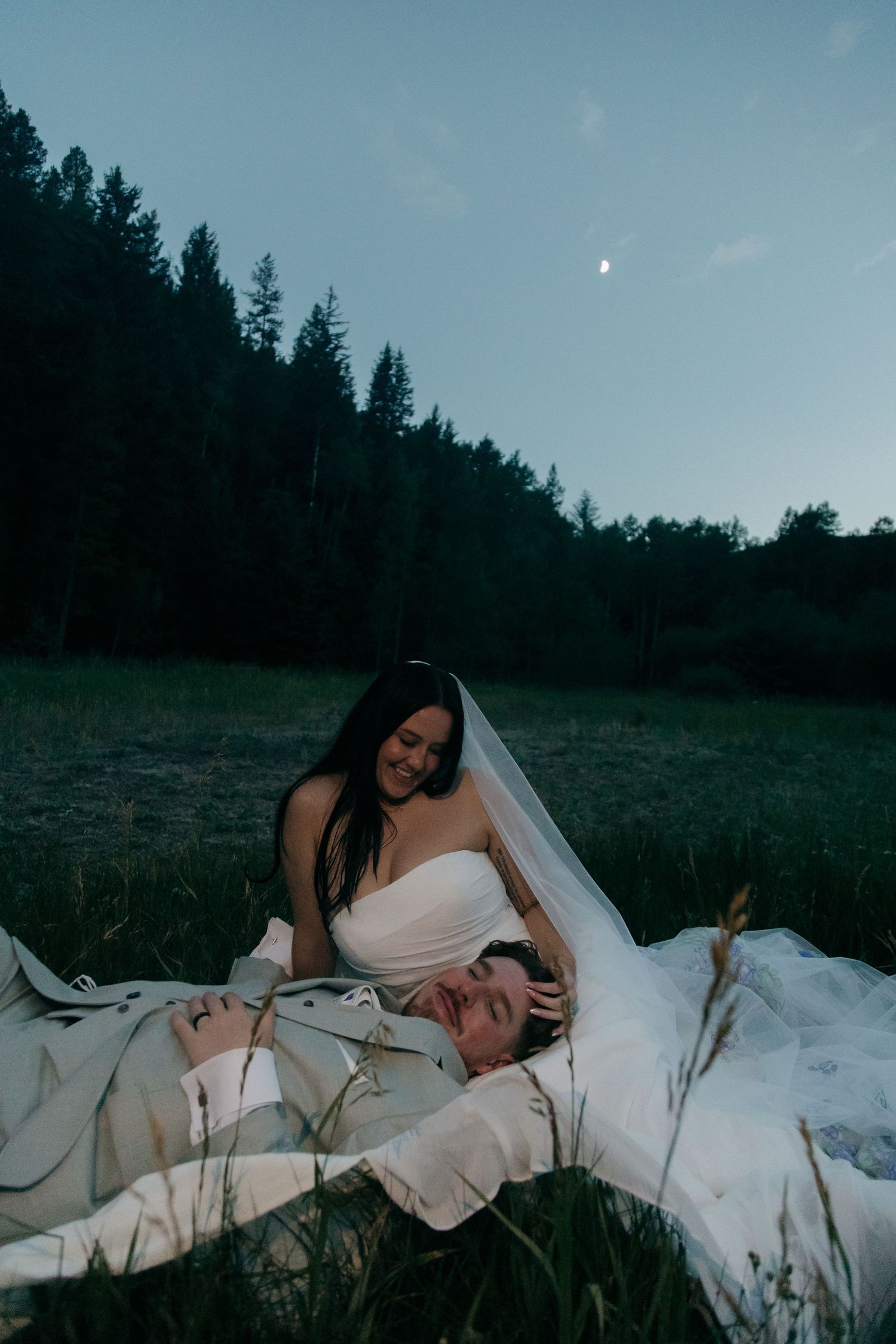 Bride in veil smiles at groom lying in her lap in moonlit forest meadow at dusk.