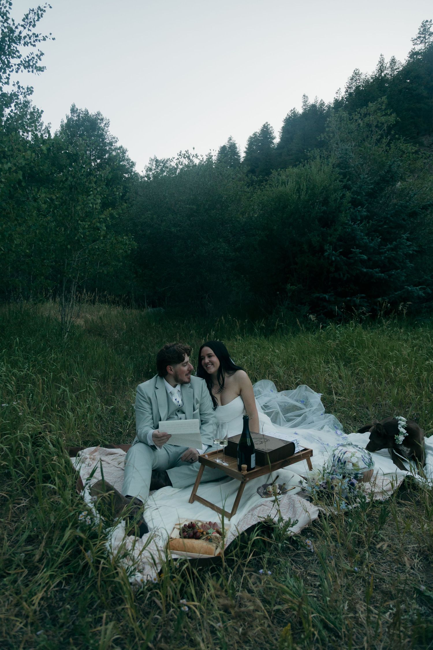 Bride and groom sitting on blanket in meadow laughing during outdoor forest elopement picnic.