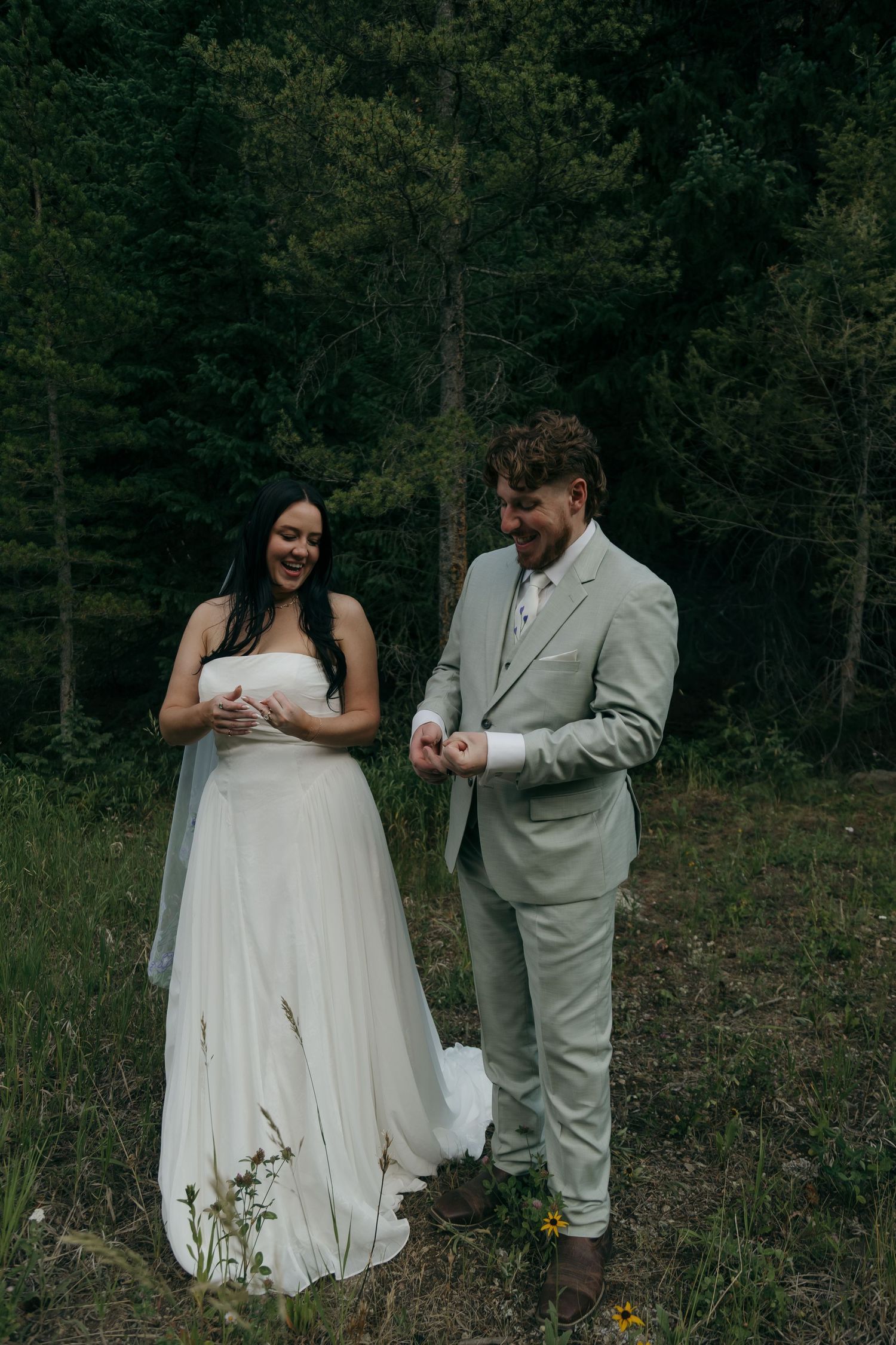 Bride and groom laugh together while exchanging rings in a lush forest wedding setting.