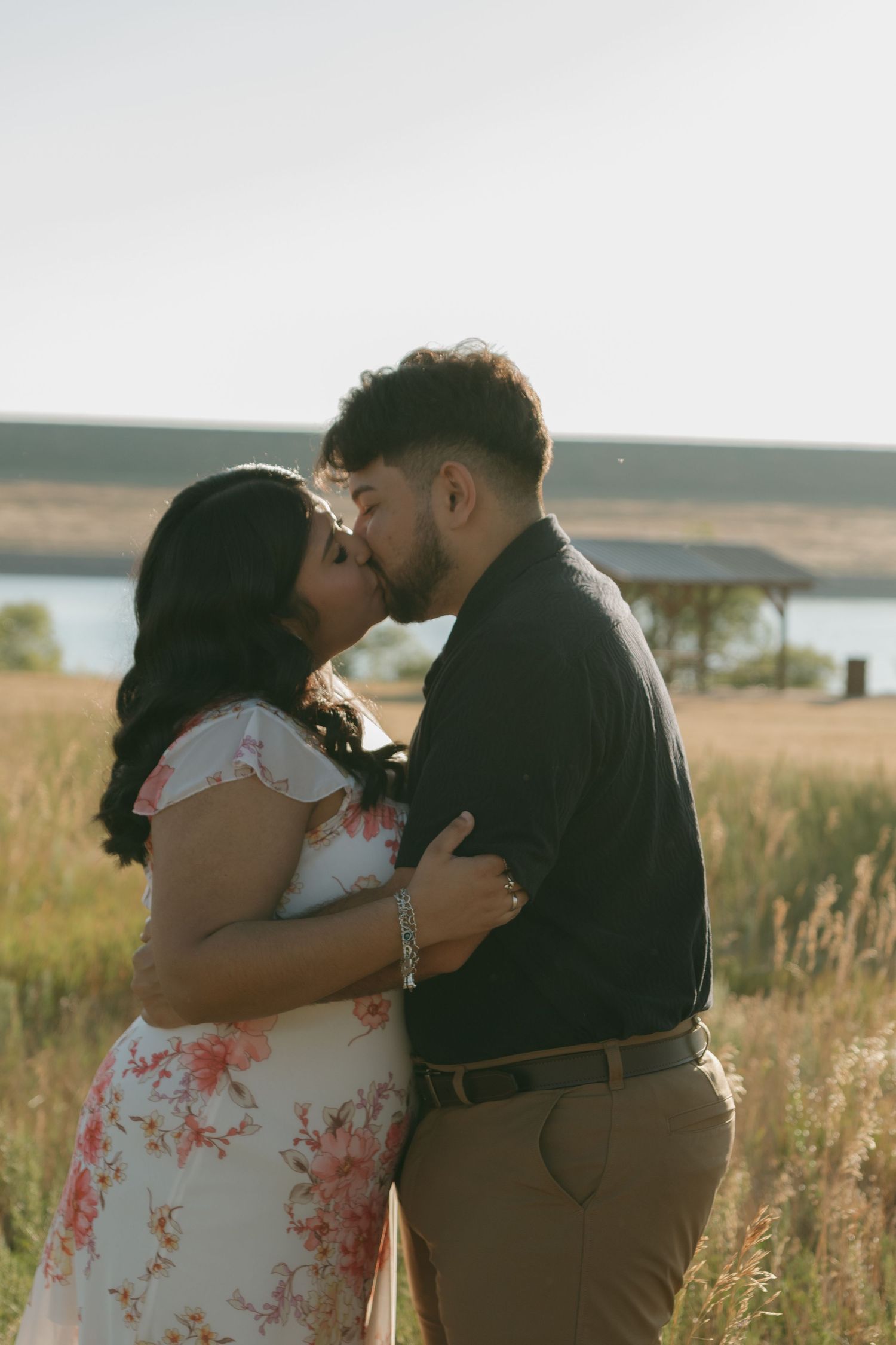 A couple shares a kiss in a scenic field with a lake and rolling hills in the background.